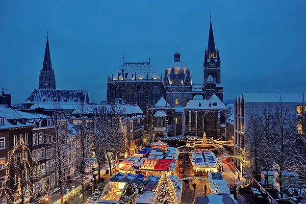 Eine Luftaufnahme des Weihnachtsmarktes in Aachen zeigt schneebedeckte Dächer und Gebäude, die den Markt mit seinen beleuchteten Buden und Weihnachtsdekorationen umgeben.