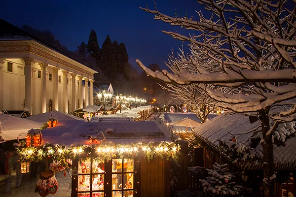  Auf dem festlichen Weihnachtsmarkt in Baden-Baden sind schneebedeckte Stände und funkelnde Lichter zu sehen. Schneebedeckte Zweige von Bäumen umrahmen die Szene.