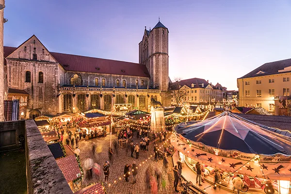 Der Weihnachtsmarkt in der Altstadt von Braunschweig, Niedersachsen, ist mit vielen beleuchteten Buden und einem Karussell im Vordergrund zu sehen. Der Dom, eine große Kirche mit einem hohen Turm, steht prominent im Hintergrund. Der Boden ist mit Pflastersteinen gepflastert, und man kann Menschen sehen, die um den Markt herumgehen. Der Himmel ist klar und wird von der untergehenden Sonne leicht angestrahlt. Dieses Bild wurde von der Braunschweig Stadtmarketing GmbH zur Verfügung gestellt.