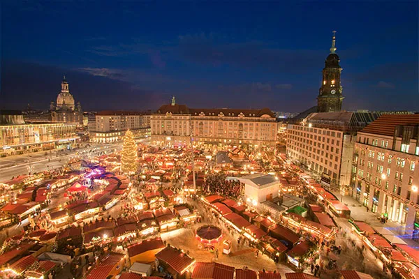 Auf dem Altmarkt in Dresden ist ein großer Weihnachtsmarkt mit vielen beleuchteten Ständen und einem hohen Weihnachtsbaum in der Mitte aufgebaut. Der Markt ist von verschiedenen Gebäuden umgeben, und die Szene wurde in den Abendstunden bei dunkelblauem Himmel aufgenommen. Bemerkenswerte Bauwerke wie die Frauenkirche und die Kreuzkirche sind im Hintergrund zu sehen.