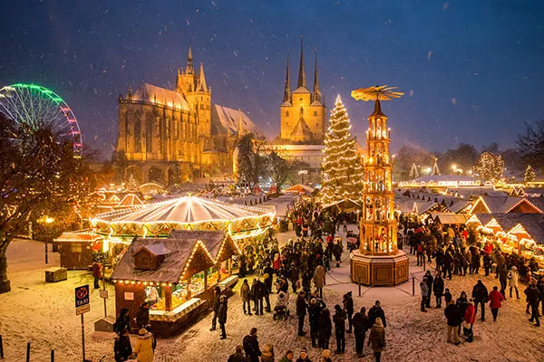 Auf dem Erfurter Weihnachtsmarkt auf dem Domplatz laufen Menschen zwischen den mit bunten Lichtern beleuchteten Ständen umher. Die Szene spielt abends, wenn Schnee fällt.