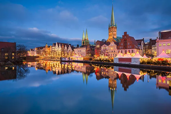 Weihnachtsmarktbuden mit festlicher Beleuchtung säumen die Trave in Lübeck. Im Hintergrund sind Kirchen mit hohen Türmen zu sehen. Der Fluss reflektiert die Lichter und Strukturen und erzeugt so einen spiegelähnlichen Effekt.