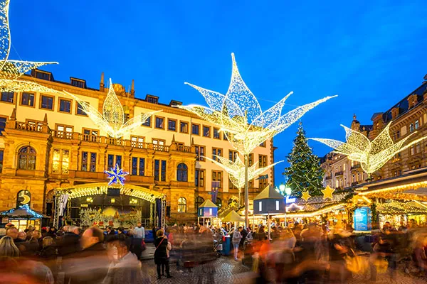 Gut besuchter Weihnachtsmarkt mit beleuchteten Sternschnuppeninstallationen vor der klassizistischen Residenzschlossfassade am Abend.