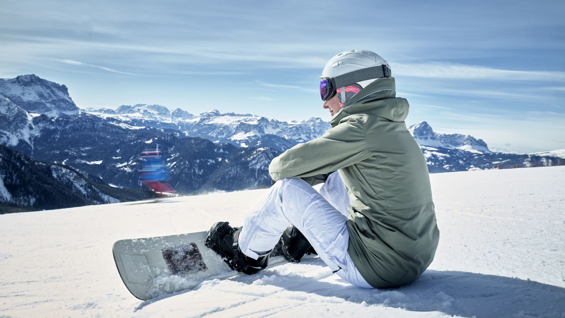 Frau sitzt mit Snowboard im Schnee und genießt die Aussicht in die Berge