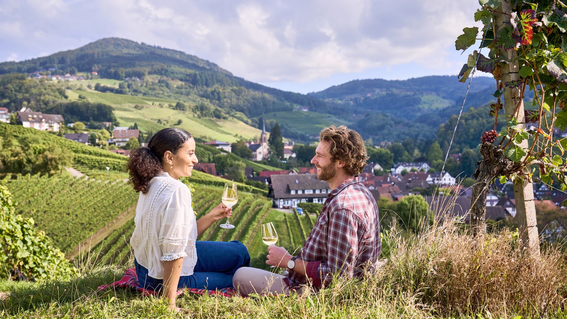 A couple sits in the middle of the vineyards of Sasbachwalden and enjoys a glass of wine with a view of the picturesque village and the surrounding hills