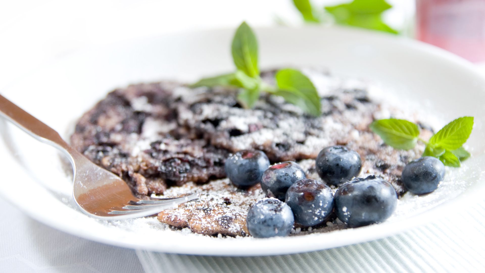 Ein Teller mit einem Heidelbeer-Pfannkuchen, bestreut mit Puderzucker. Daneben liegen frische Heidelbeeren und einige Minzblätter zur Dekoration. Eine Gabel liegt am Rand des Tellers.