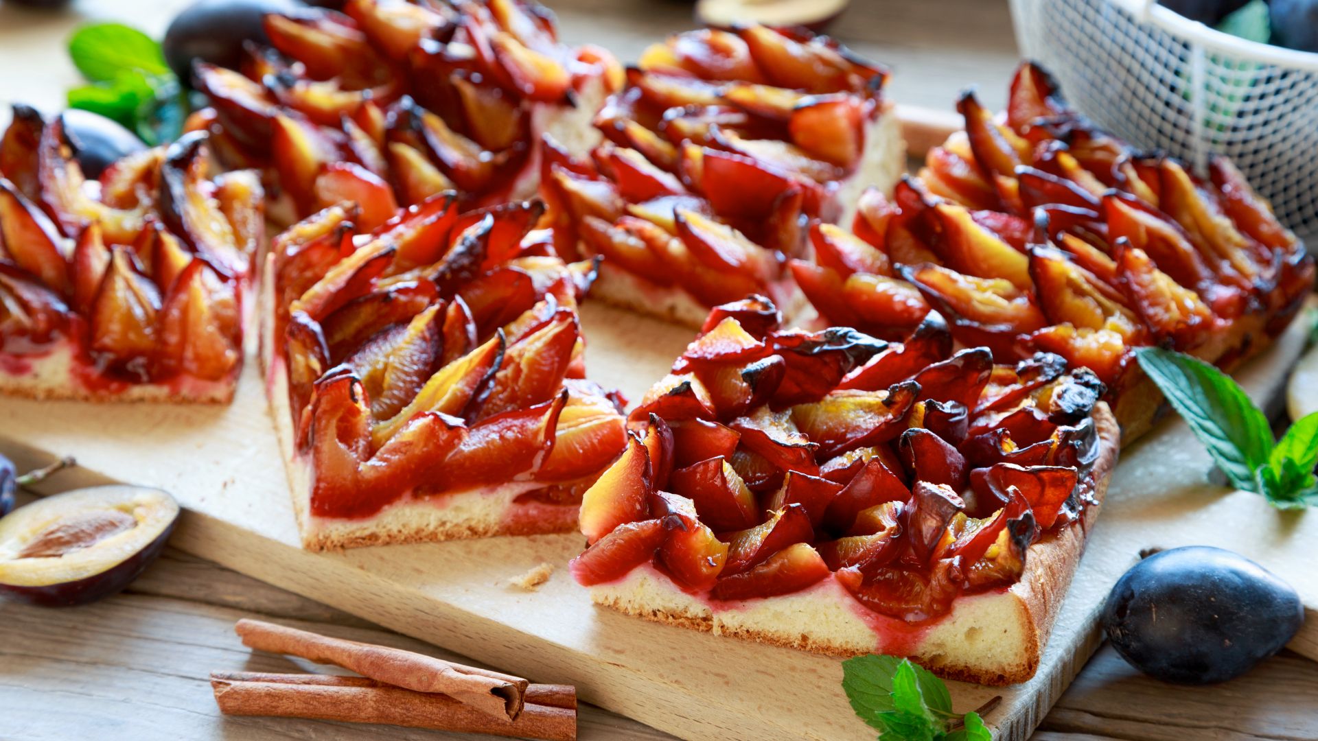 Zwetschgenkuchen, ein traditioneller deutscher Pflaumenkuchen, der in quadratische Stücke geschnitten wird, liegen auf einem Schneidebrett aus Holz, daneben frische Pflaumen und Zimtstangen.