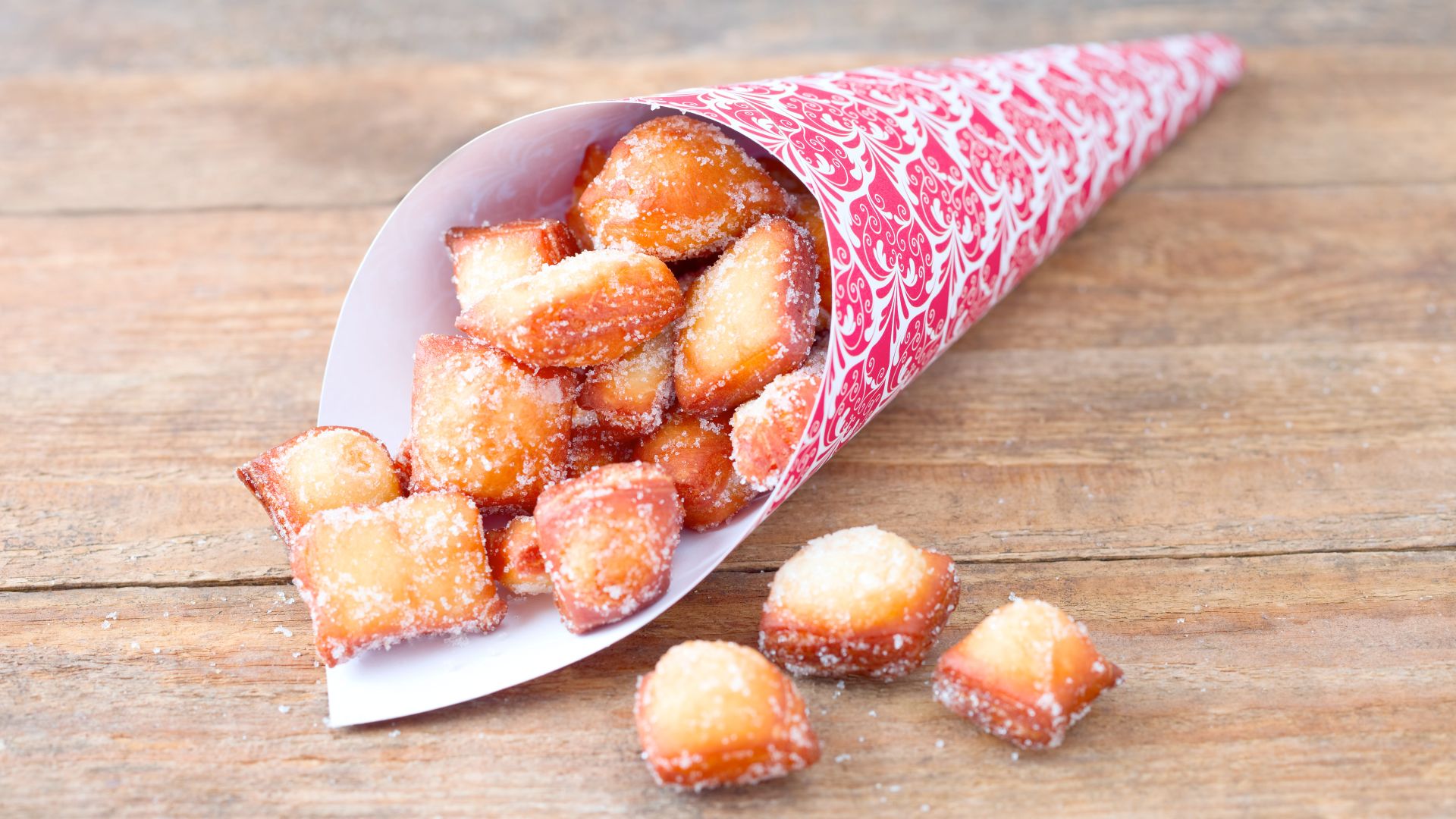 Eine Tüte mit rot-weißem Muster enthält kleine, gebackene Teigstücke, die mit Zucker bestreut sind. Einige der Teigstücke sind auf einem Holztisch verstreut. Das Bild zeigt keine erkennbaren Orte oder Gebäude.