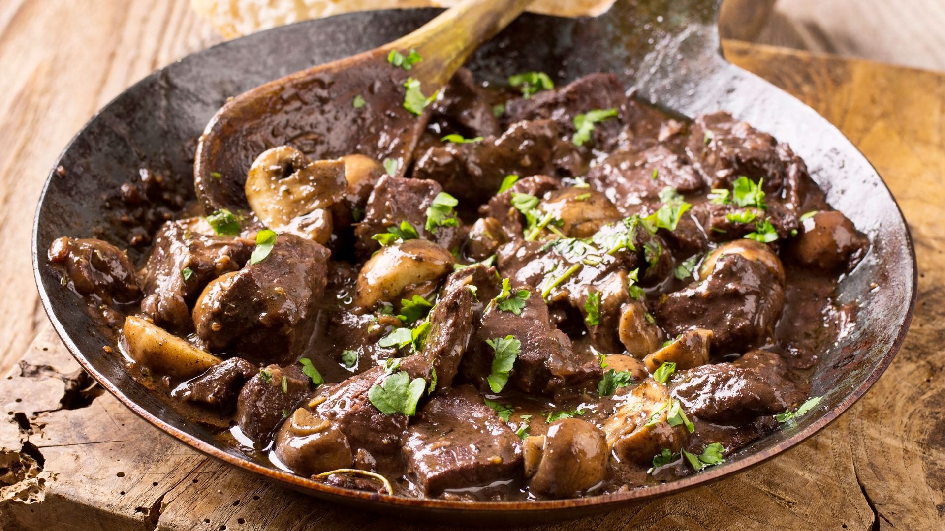 Eine dunkle Pfanne mit einem Gericht aus braunem Fleisch, Pilzen und grünen Kräutern, serviert auf einem Holzbrett. Ein Holzlöffel liegt in der Pfanne, und im Hintergrund ist ein Stück Brot sichtbar. Das Gericht ist mit frischen Kräutern garniert.