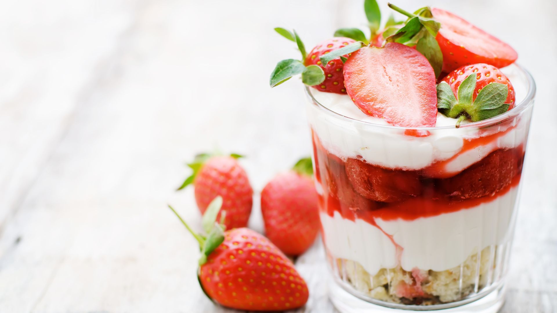 Ein Glas mit einer Dessertschichtung aus Sahne, Erdbeersauce und frischen Erdbeeren, dekoriert mit halbierten Erdbeeren, steht auf einem hellen Holztisch. Im Hintergrund liegen weitere frische Erdbeeren. Das Dessert wirkt frisch und farbenfroh.