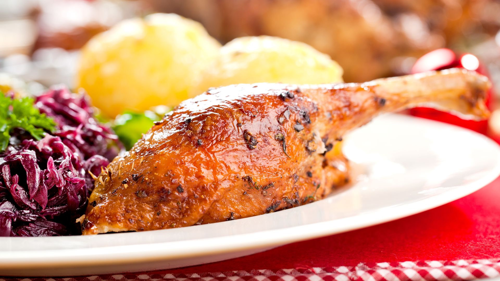 Ein Teller mit einem gebratenen Entenbein, begleitet von Rotkohl und Kartoffelknödeln. Das Essen ist dekorativ angerichtet, und im Hintergrund sind weitere verschwommene Elemente eines gedeckten Tisches zu erkennen. Ein rotes Tischtuch und ein karierter Stoffsaum sind unter dem Teller sichtbar.