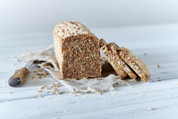 Ein Laib rheinisches Schwarzbrot und drei Scheiben davon liegen auf einem Tuch auf einem hellen Holztisch, daneben liegt ein Messer mit Holzgriff