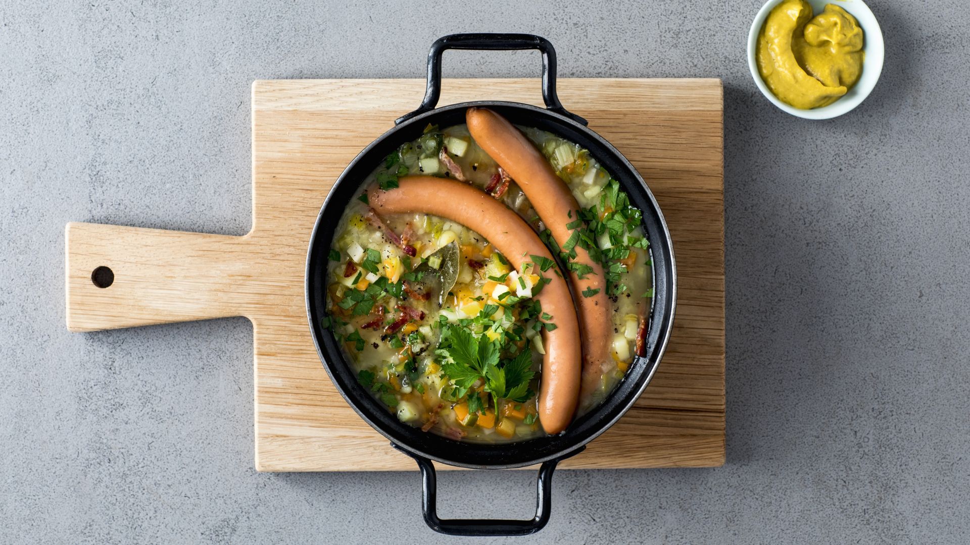 Eine Schüssel Berliner Kartoffelsuppe mit zwei Wiener Würstchen steht auf einem hölzernen Schneidebrett