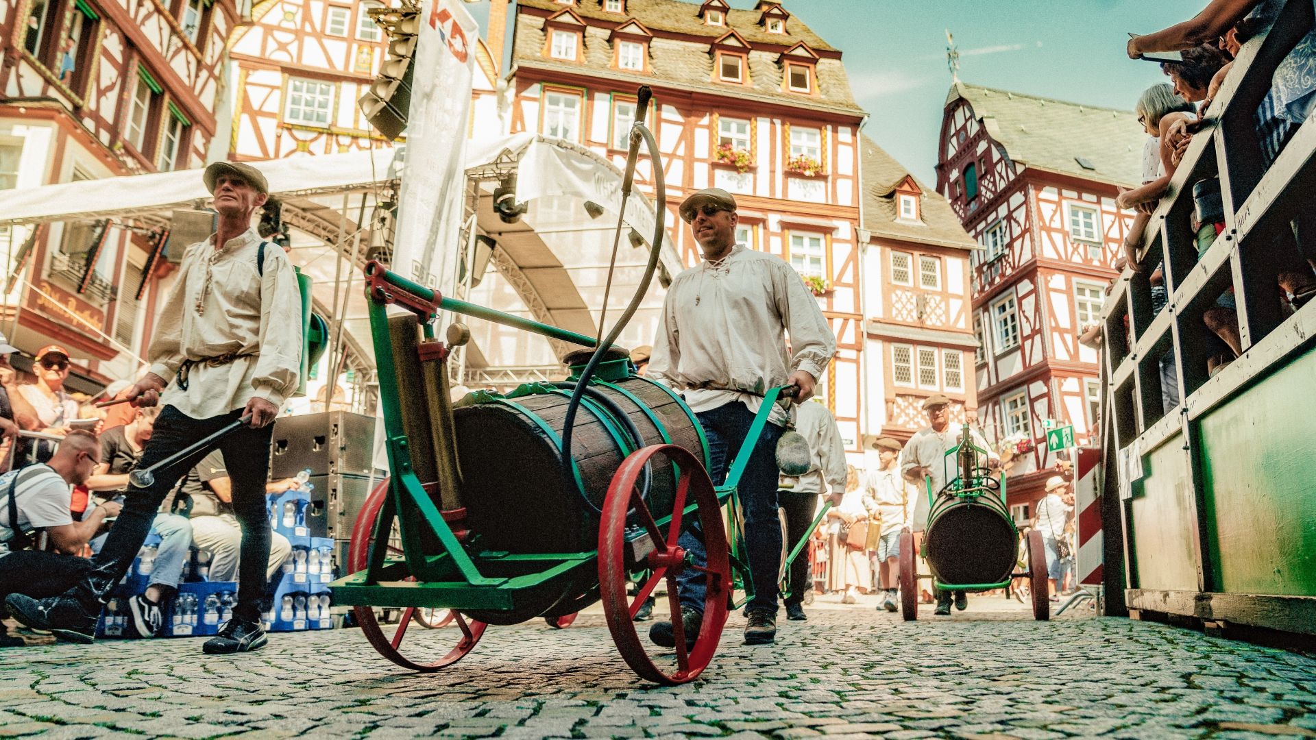 Fête historique dans une vieille ville avec des maisons à colombages, où les participants en costumes traditionnels tirent des tonneaux en bois sur des charrettes à bras à travers les rues.