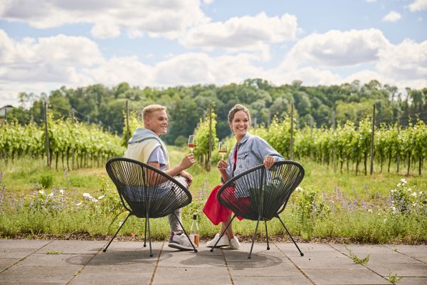 Ein Mann mit einer Beinprothese und eine Frau sitzen auf schwarzen Stühlen vor einem Weinberg in Vogtsburg im Kaiserstuhl. Sie genießen den Wein aus dem Hochschwarzwald.