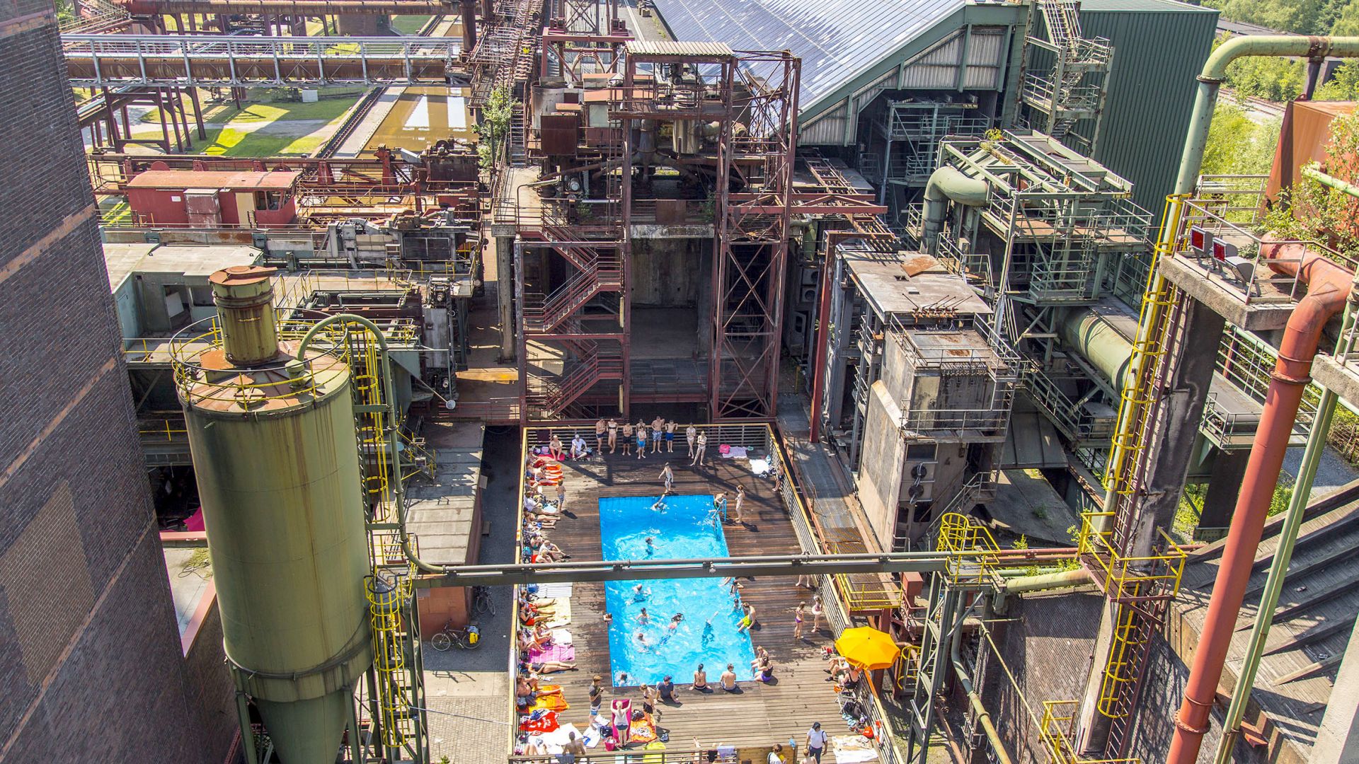 Große Stahlkonstruktionen der Zeche Zollverein in Essen umgeben ein Freibad, das Werksschwimmbad, in dem viele Menschen schwimmen und sich sonnen