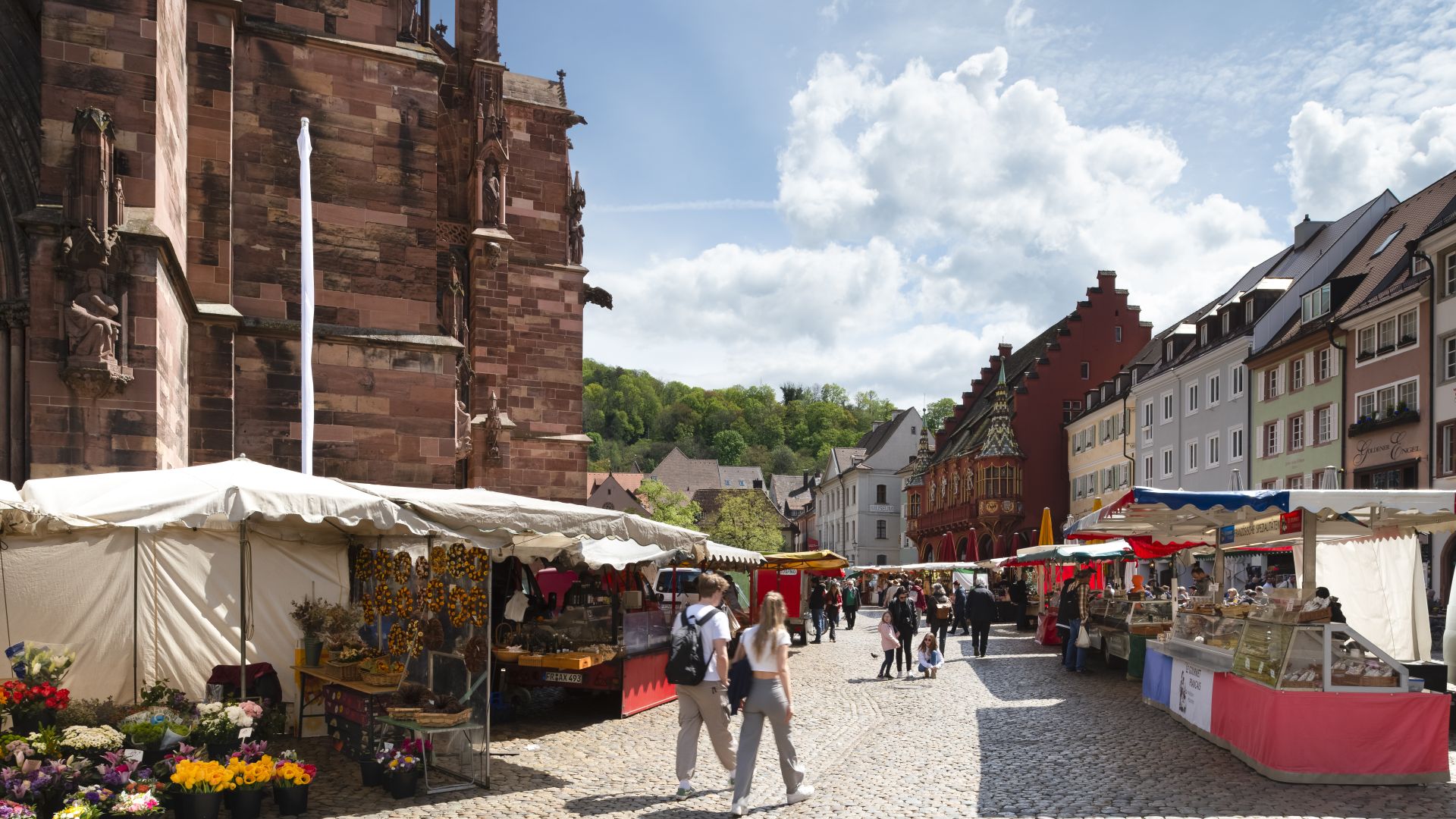Auf einer Kopfsteinpflasterstraße in Freiburg findet der Münstermarkt statt, mit Marktständen auf beiden Seiten. Die Menschen gehen zwischen den Ständen umher. Die linke Seite zeigt einen Teil eines roten Steingebäudes. Im Hintergrund sind mehrere bunte Gebäude und ein grüner Hügel zu sehen.