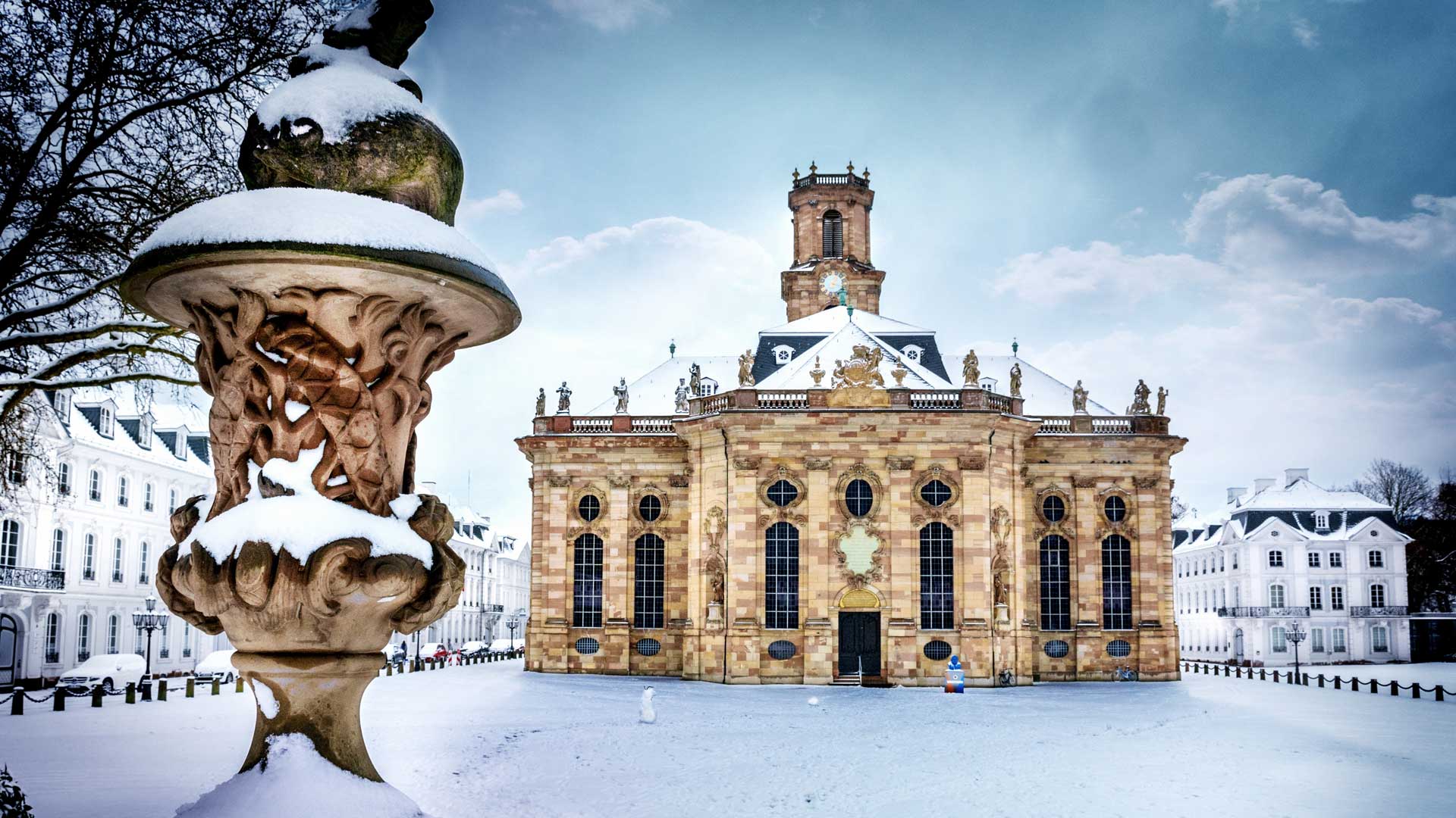 Die Ludwigskirche in Saarbrücken, Saarland, ist mit Schnee bedeckt. Die Kirche hat große Fenster und Statuen auf dem Dach. Im Vordergrund ist ein Steinornament mit Schnee darauf zu sehen. Der Boden und die umliegenden Gebäude sind ebenfalls mit Schnee bedeckt. Der Himmel ist klar mit einigen Wolken.