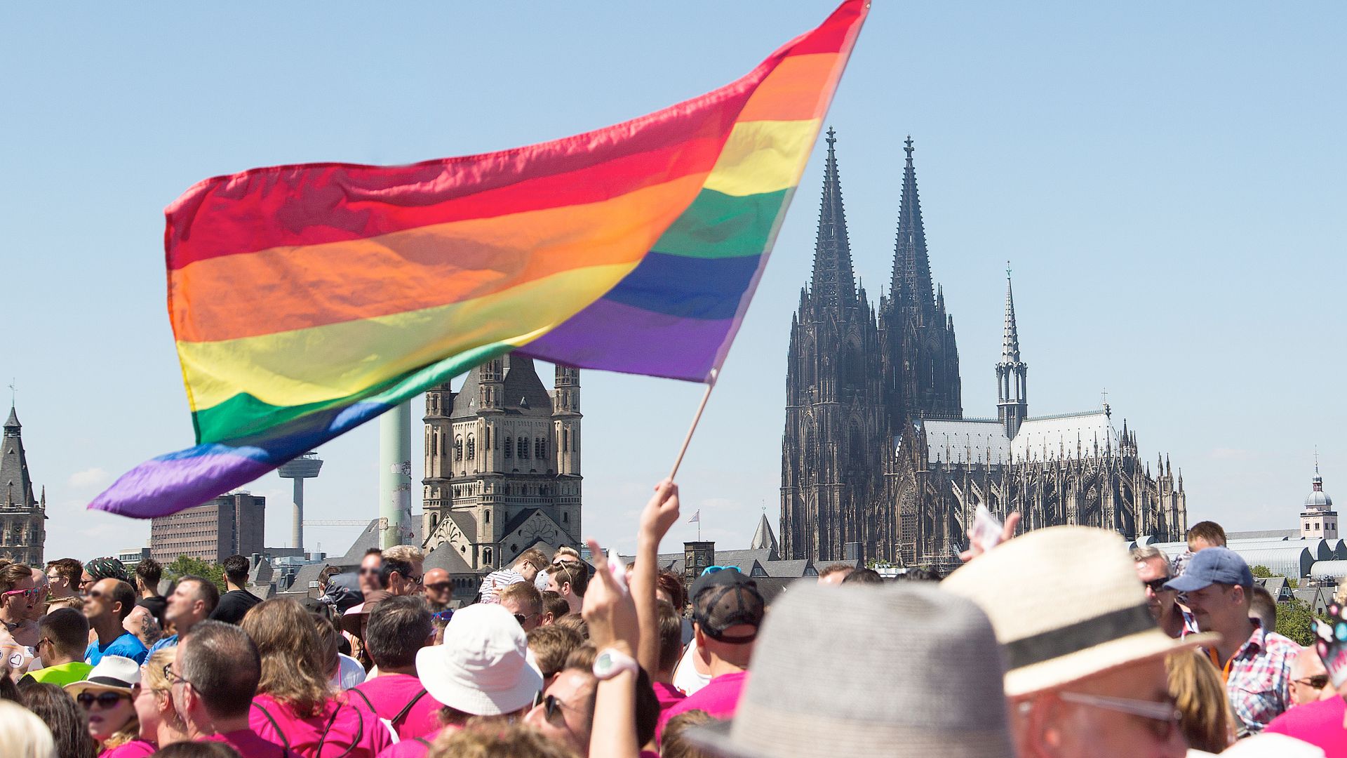 Eine Menschenmenge mit pinken T-Shirts trägt eine Regenbogenfahne vor sich. Im Hintergrund ist der Kölner Dom in Köln klar erkennbar. Der Himmel ist blau und wolkenlos.