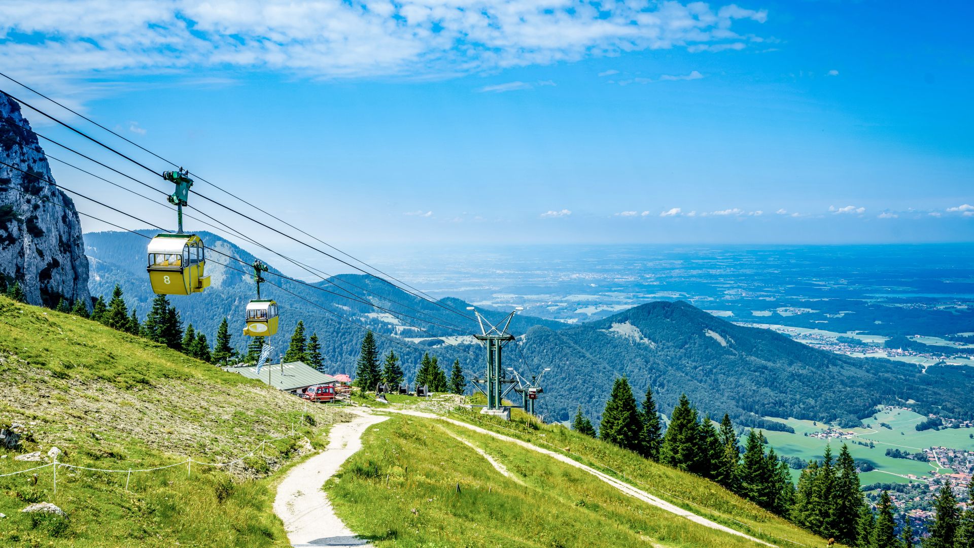 Eine gelbe Seilbahn mit mehreren Gondeln fährt über eine grüne Gebirgslandschaft. Ein schmaler Wanderweg schlängelt sich den Berg hinauf, flankiert von Tannenbäumen und einer felsigen Steilküste links im Bild. Im Hintergrund erstreckt sich eine weite Landschaft unter blauem Himmel mit vereinzelten Wolken.