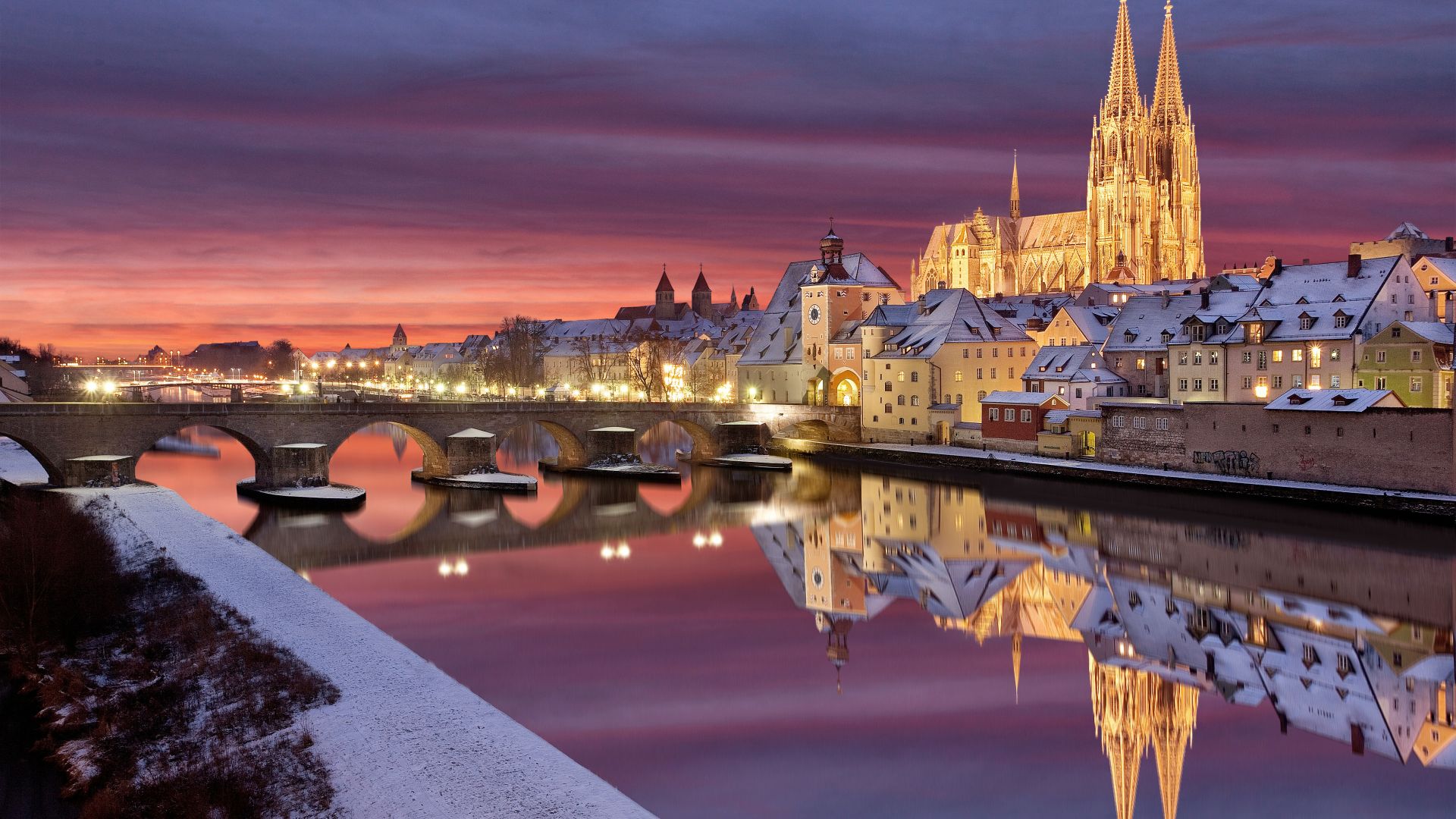 In Regensburg, Bayern, führt die steinerne Brücke über einen Fluss, der in die Altstadt führt. Der Dom St. Peter steht hoch und beleuchtet im Hintergrund. Die Gebäude entlang des Flussufers sind mit Schnee bedeckt, der sich im ruhigen Wasser spiegelt. Der Himmel weist rote und blaue Farbtöne auf, was darauf hindeutet, dass es entweder Morgengrauen oder Abenddämmerung ist. Der schneebedeckte Boden und die beleuchteten Fenster der Gebäude tragen zu der heiteren Atmosphäre bei.