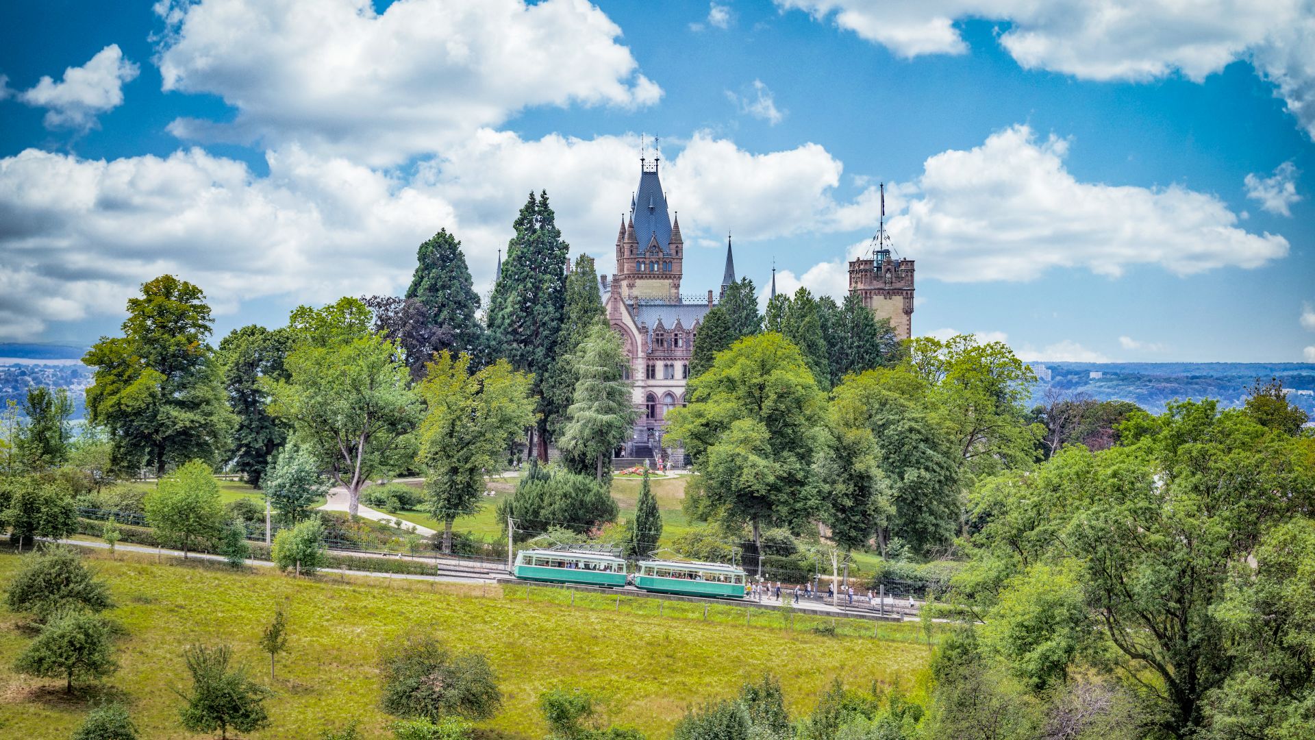 Die Burg Drachenfels mit einem hohen Turm und markanten Giebeln steht umgeben von zahlreichen Bäumen, darunter sind Spazierwege erkennbar. Ein grüner Zug fährt auf Gleisen im Vordergrund entlang, während im Hintergrund eine hügelige Landschaft zu sehen ist. Der Himmel ist bewölkt mit vereinzeltem Sonnenschein.