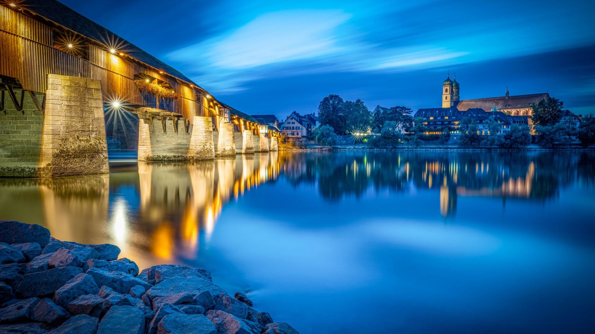 The illuminated wooden bridge stretches across the Rhine in Bad Säckingen in a night-time scene. To the right of the bridge, buildings and a church tower can be seen reflected in the water. The sky is deep blue with a few clouds.