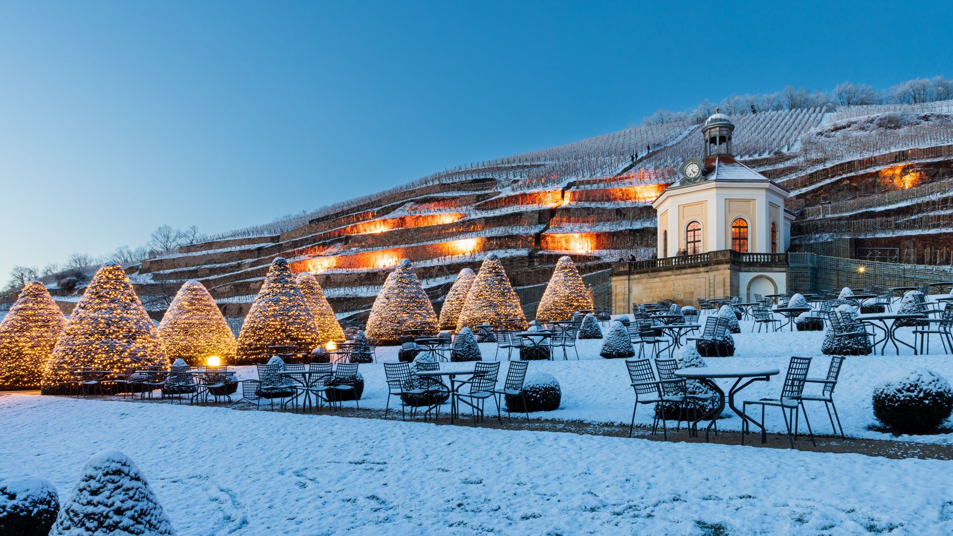 Schnee bedeckt den Boden und die Stühle in einem Park von Schloss Wackerbarth in Radebeul. Kegelförmige Bäume mit Lichtern säumen das Gelände, und an den Hängen sind Weinberge zu sehen. Auf der rechten Seite steht ein Gebäude mit einem Uhrenturm.