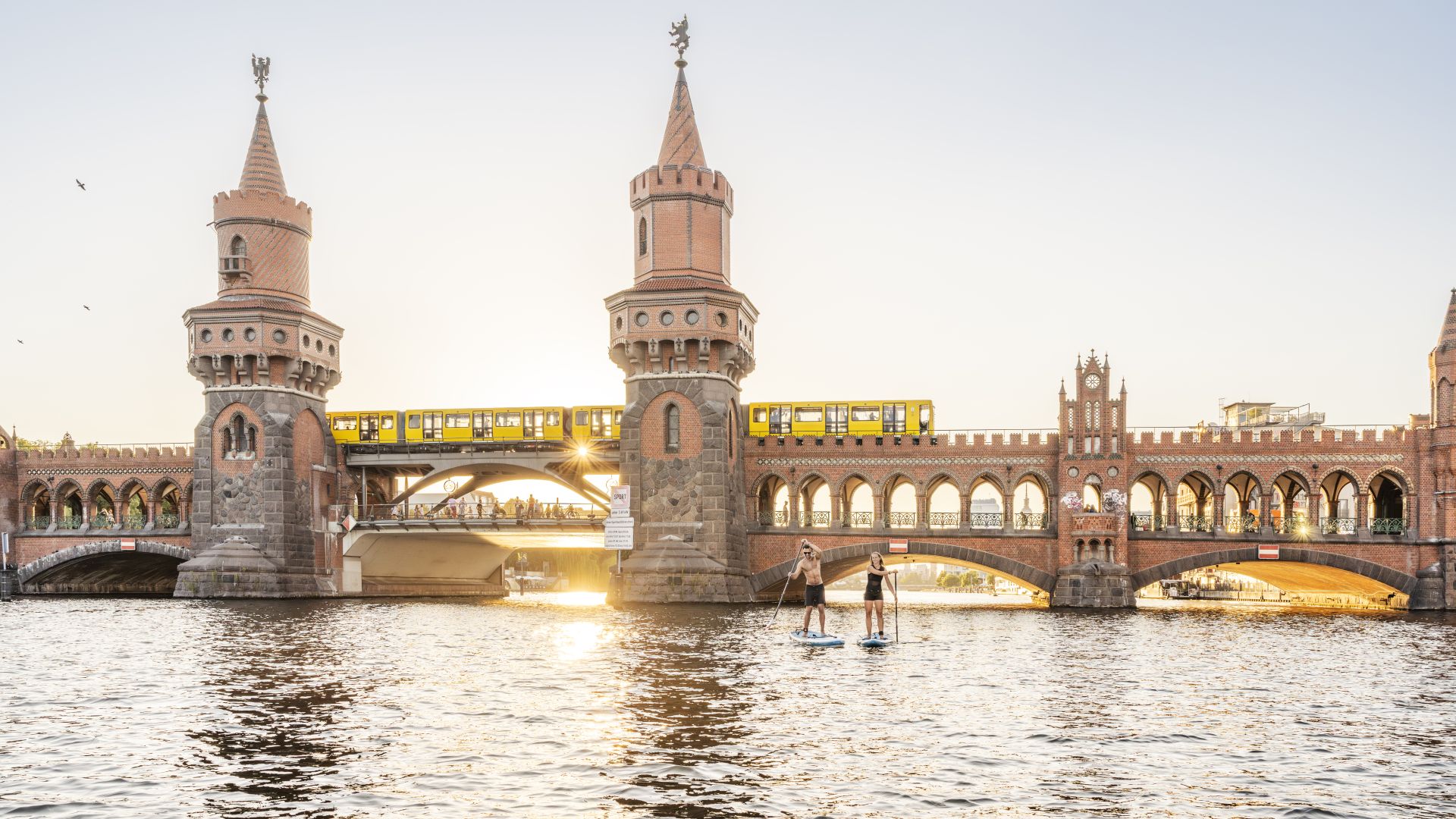 A young woman and a man go stand-up paddleboarding on the Spree in front of the Oberbaumbrücke as the sun sets.
