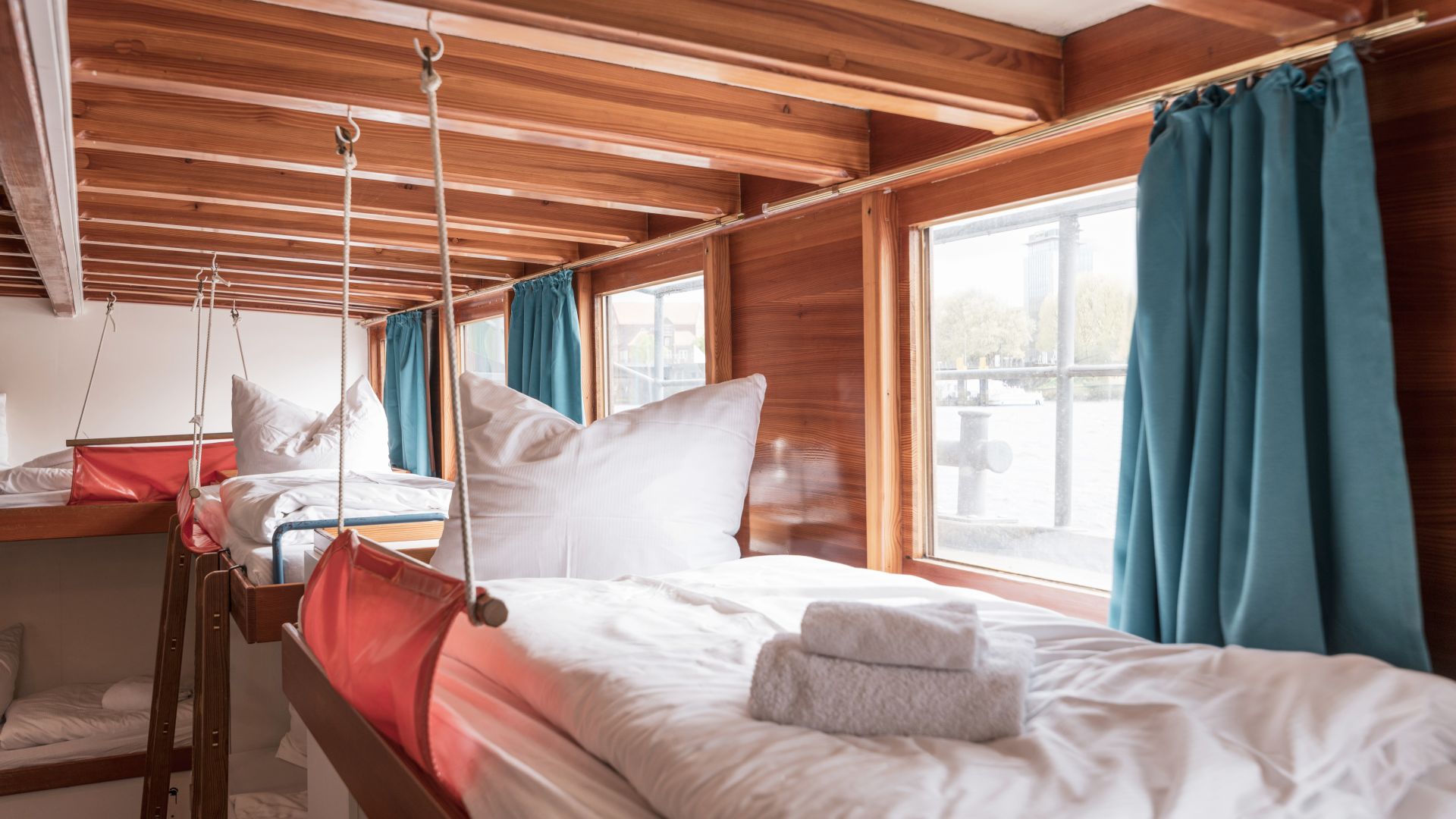 Innenraum eines Schiffs mit mehreren Schlafkojen aus Holz, die entlang großer Fenster angeordnet sind. Kojen sind mit weißen Decken und Kissen ausgestattet, blaue Vorhänge sind an den Fenstern angebracht. Durch die Fenster ist Wasser und ein Teil der Uferpromenade zu sehen.