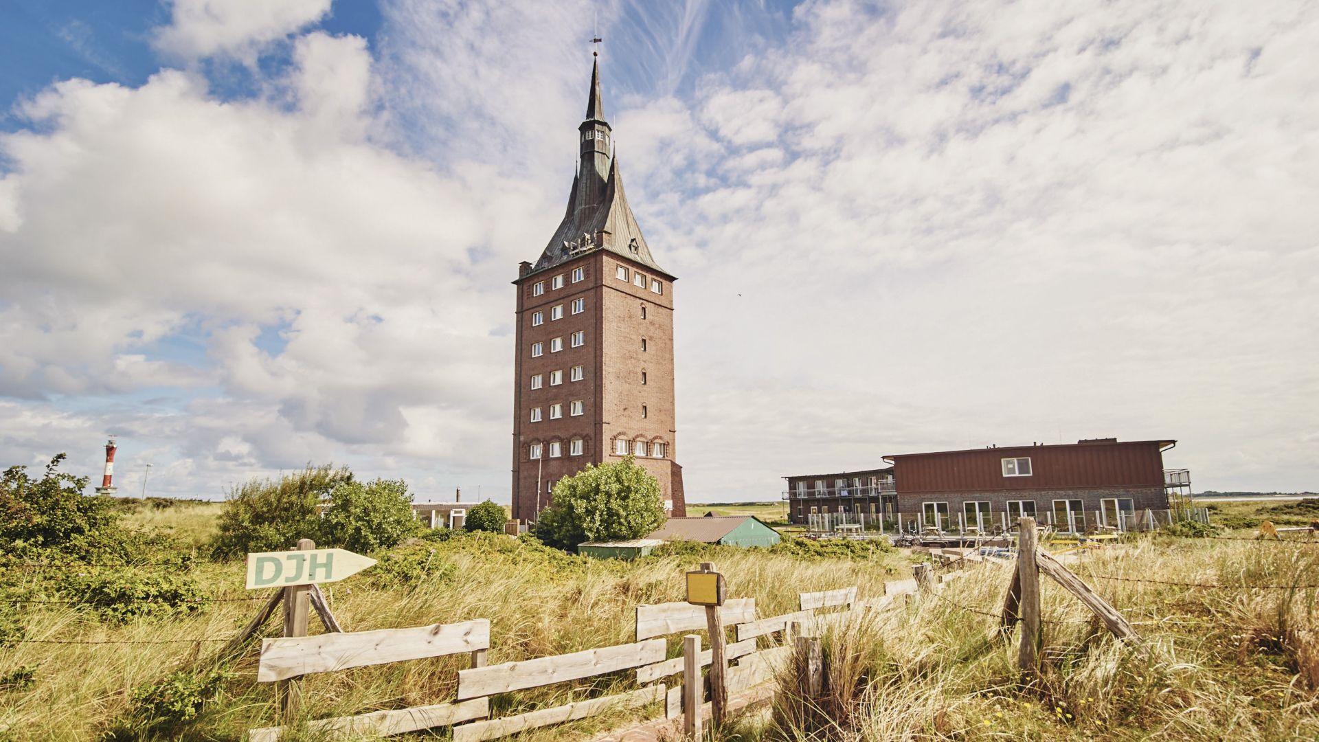 Ein Turm mit spitzem Dach erhebt sich in einer flachen Landschaft, umgeben von Gras und einem Holzzaun im Vordergrund. Im Hintergrund ist ein Gebäude mit moderner Architektur und ein Leuchtturm zu sehen. Ein Schild mit der Aufschrift „DJH“ weist auf eine Jugendherberge hin.