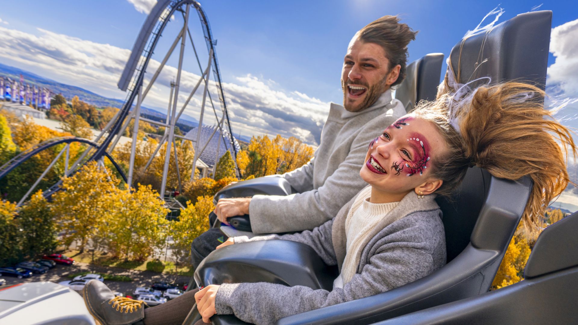 Mann und Mädchen mit Halloween-Makeup sitzen fröhlich, mit wehenden Haaren in einer fahrenden Achterbahn.