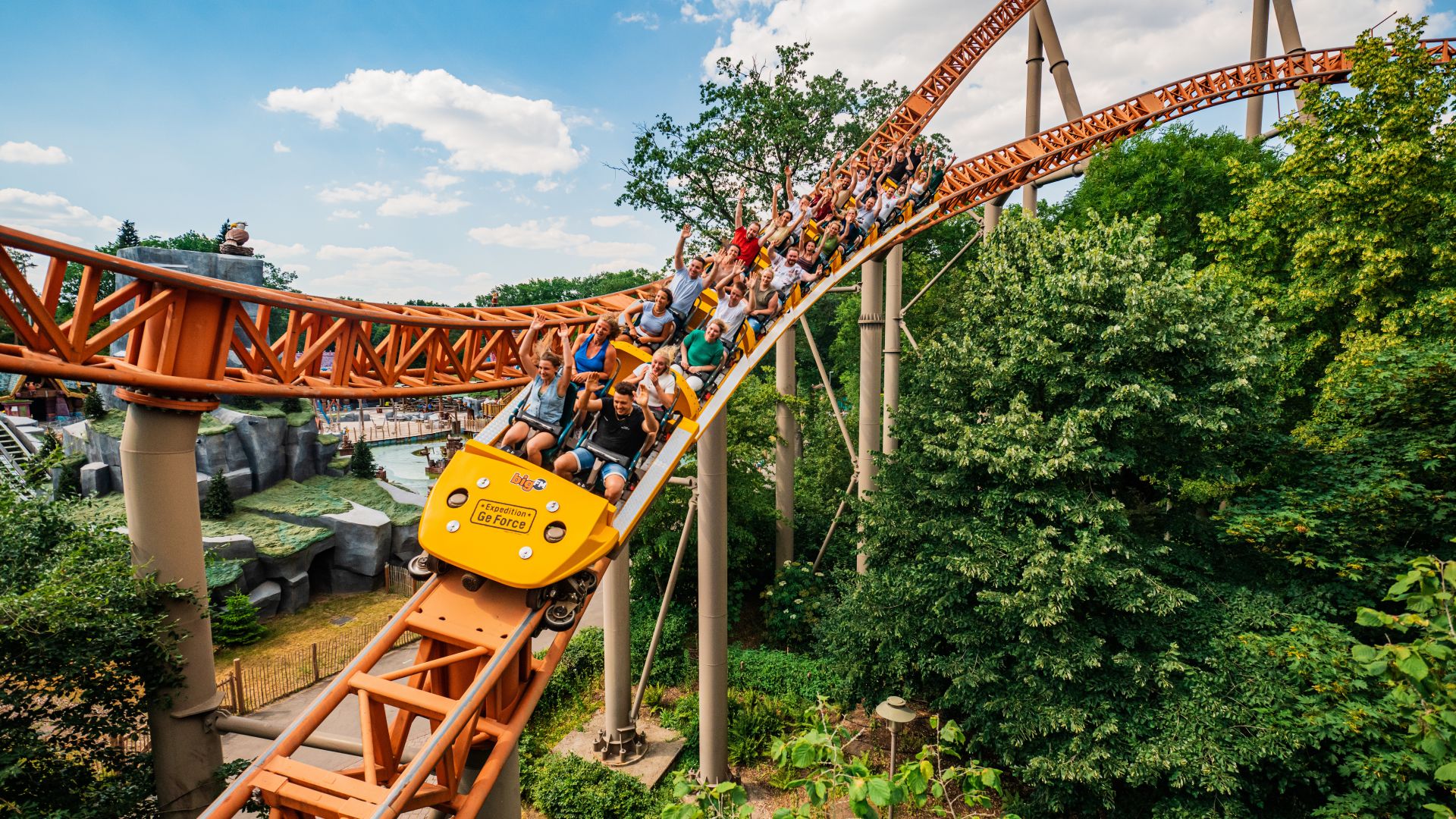 voll besetzter Achterbahnzug einer orangefarbenen Stahlachterbahn zwischen grünen Bäumen