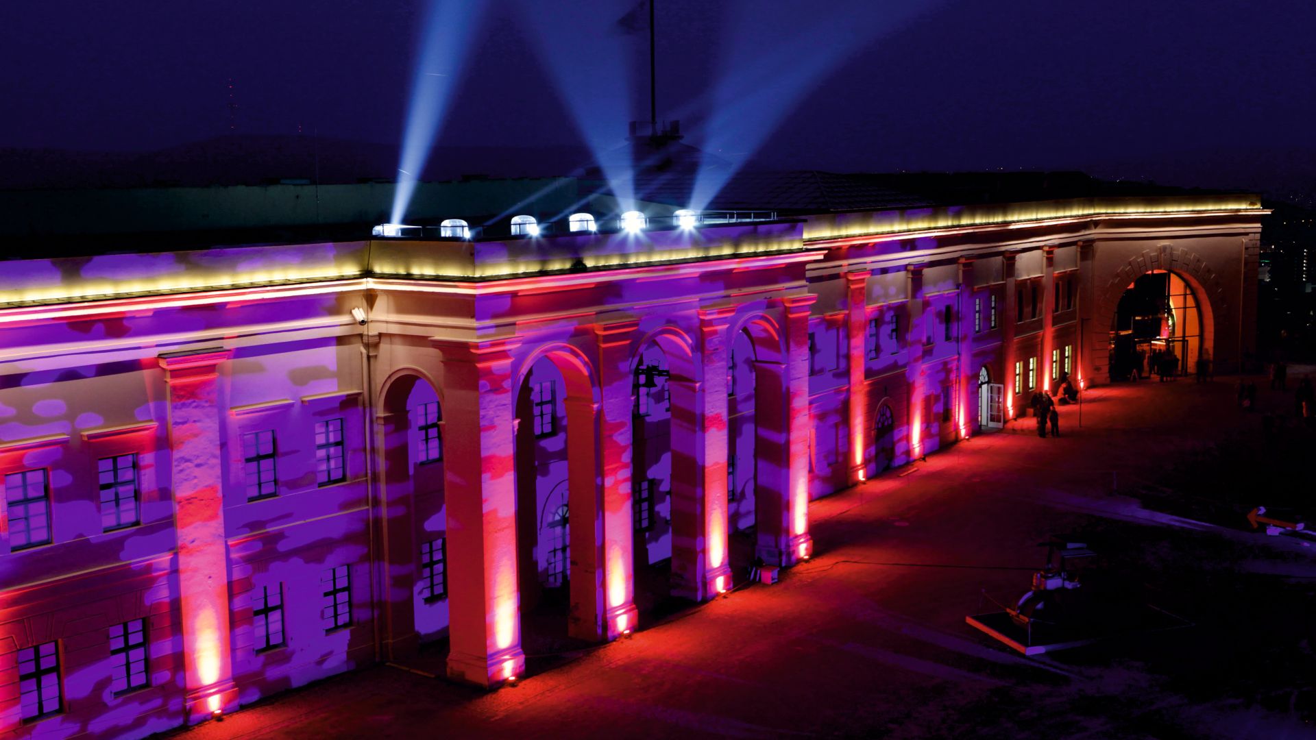 A large building with arched doorways and windows. It is illuminated at night with colorful lights, mainly in red and purple, and several spotlights can be seen on the roof. The building appears to be a fortress or historical structure, and fine patterns are projected onto the façade. Some people can be seen near the entrance to the building. The scene takes place at night in Koblenz, Rhineland-Palatinate.