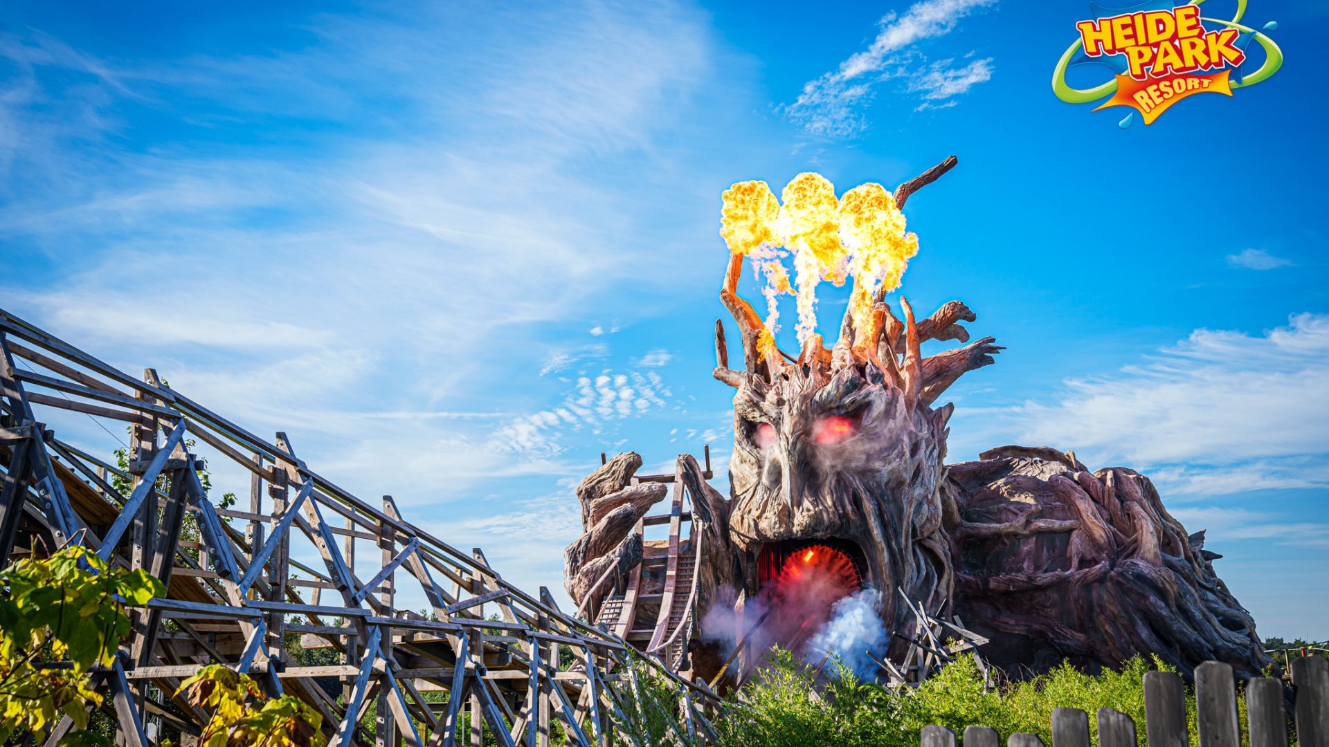 AUs dem Kopf eines furchterregenden Baummonsters steigen leuchtende Flammen empor. Im Vordergrund verläuft eine Holzachterbahn.