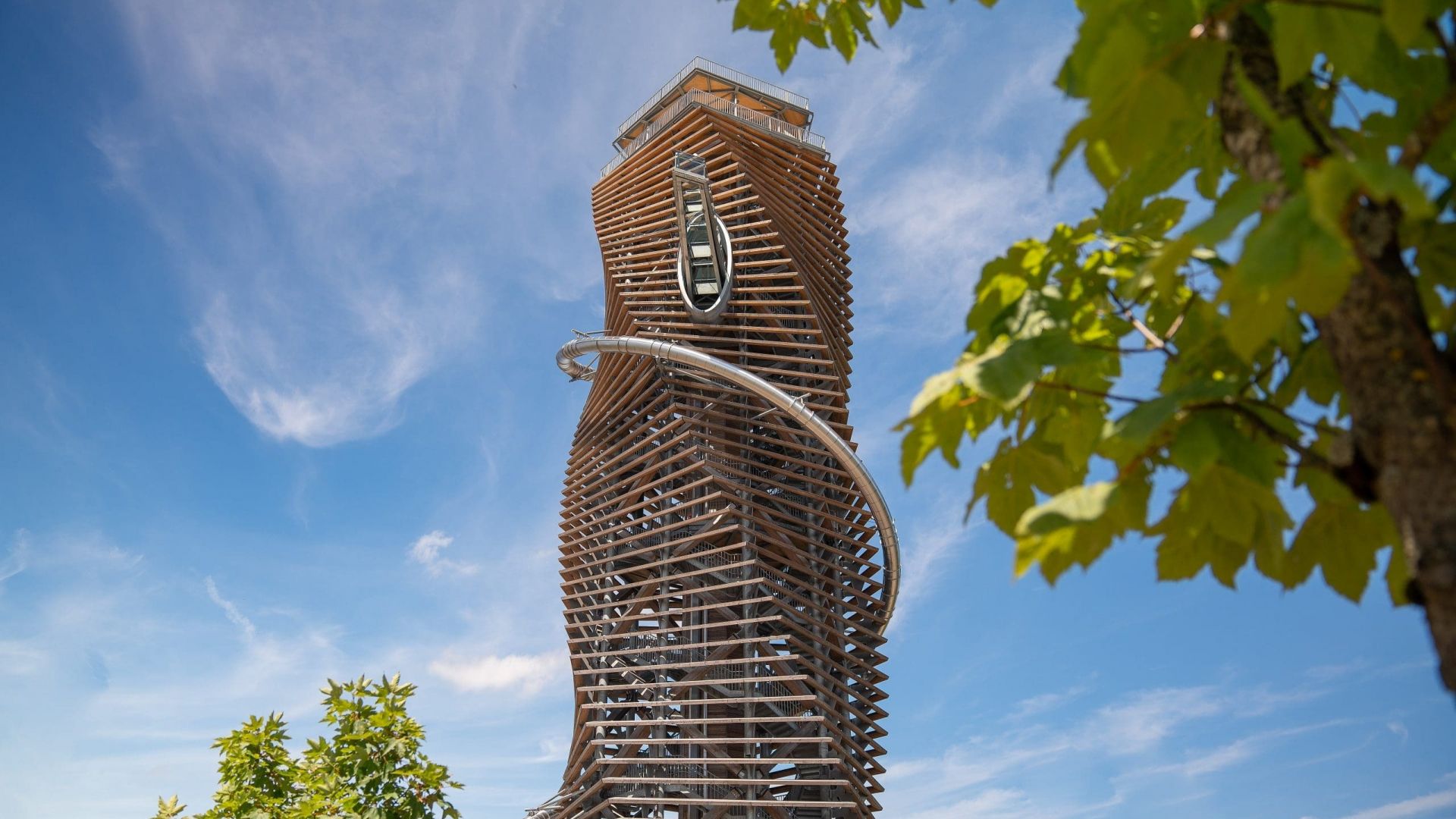 Turm mit einer modernen Holzstruktur und einer markanten, sich windenden Rutsche, die das Gebäude umhüllt, vor einem klaren Himmel und grünen Bäumen
