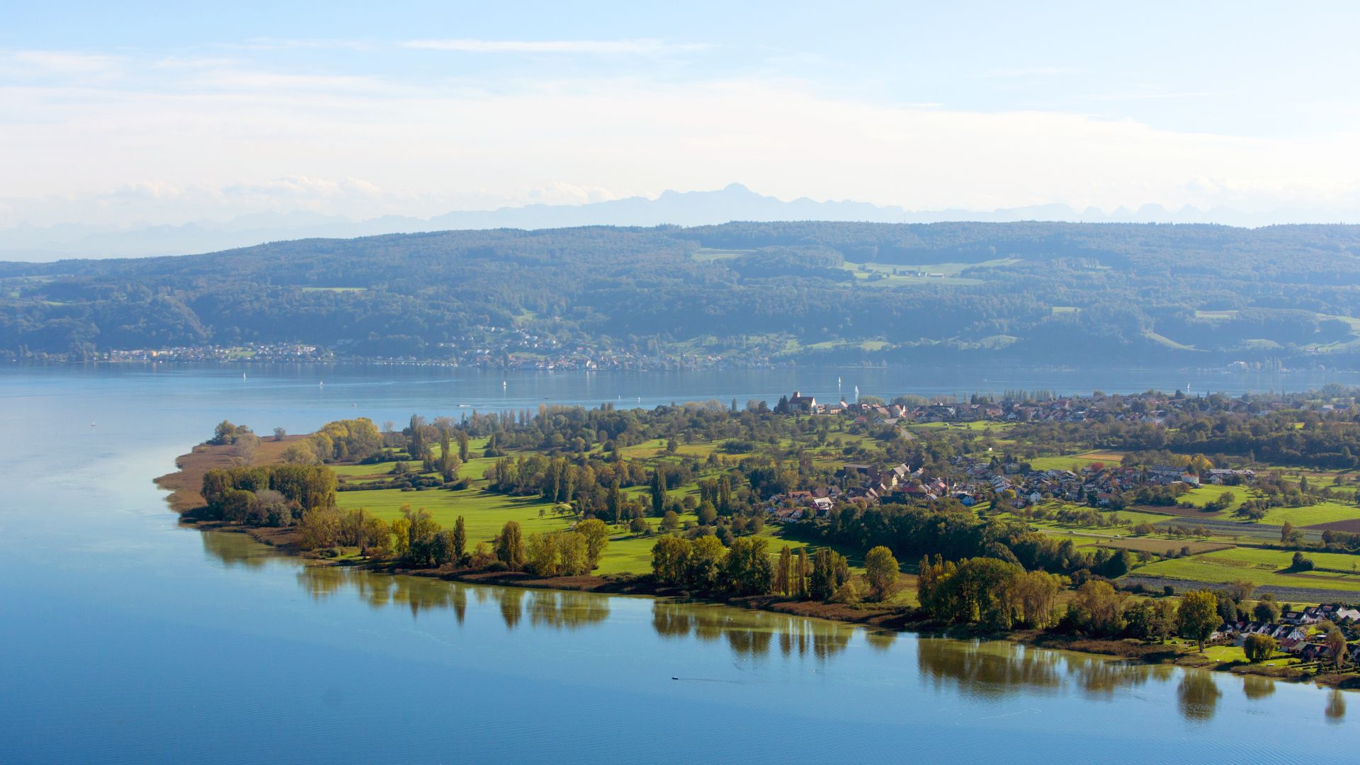 A peninsula with green fields and trees flows into a lake. Small towns and scattered buildings can be seen near the shore. The lake is calm and reflects parts of the landscape.