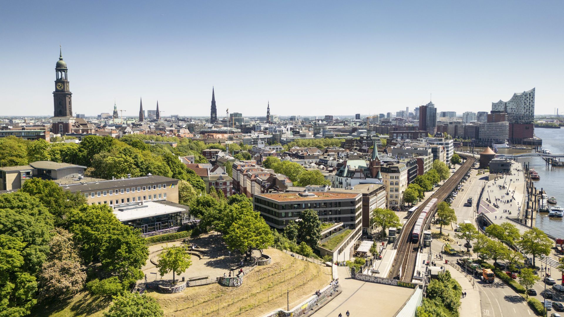 Stadtansicht von Hamburg mit dem Turm des Hamburger Michels links und der Elbphilharmonie rechts im Hintergrund. Im Vordergrund sind grüne Parkanlagen und Gebäude zu sehen. Ein Zug fährt auf oberirdischen Gleisen entlang der Elbe.