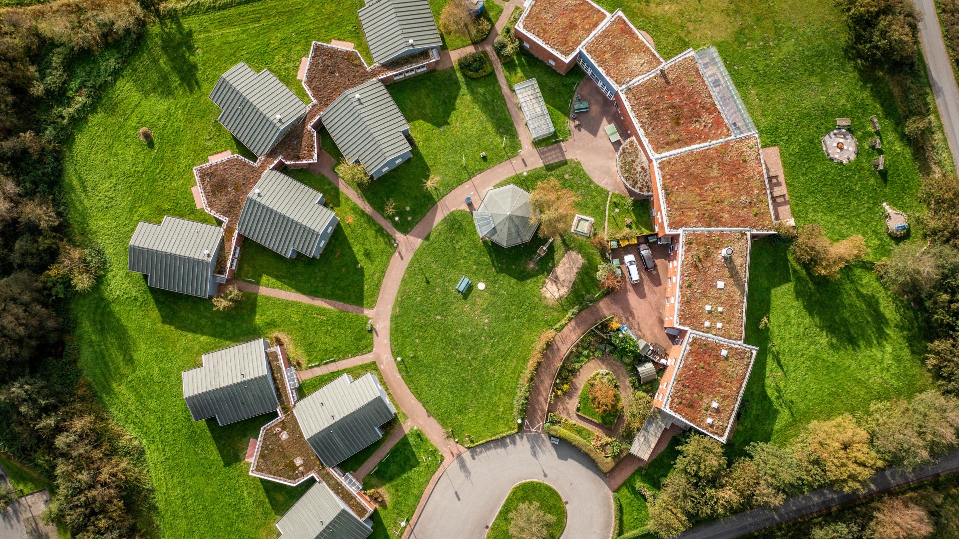 Luftaufnahme einer Anlage von Gebäuden mit begrünten Dächern, die um einen zentralen, runden Platz mit einem Pavillon angeordnet sind. Die Umgebung ist von grünen Rasenflächen und Bäumen geprägt. Eine Straße verläuft am unteren Bildrand, rechts ein Kreisverkehr.