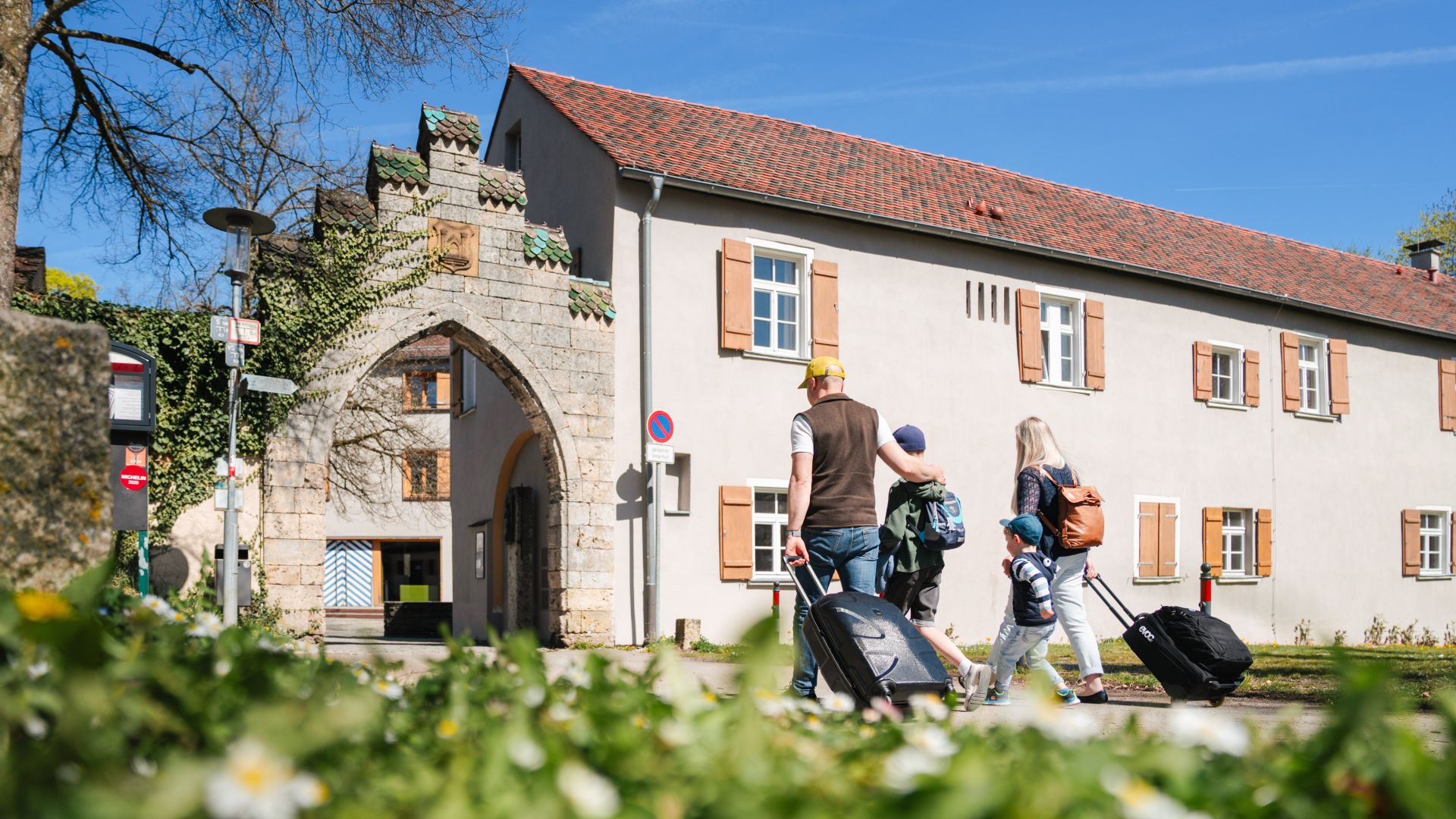 Zwei Erwachsene und zwei Kinder ziehen Koffer entlang eines Wegs vor einem historischen Steingebäude mit einem Bogentor und roten Dachziegeln. Das Gebäude wird von Efeu berankt und befindet sich in Ravensburg.