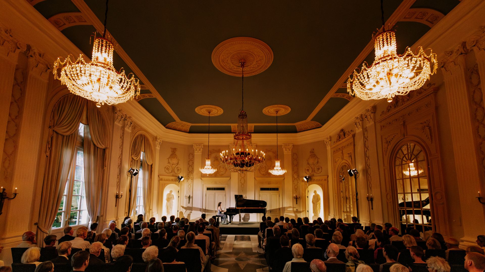 La imagen muestra una sala de conciertos durante el Festival de Beethoven en Bonn. Un pianista está actuando frente a una sala llena adornada con magníficas lámparas de araña y una decoración elegante.