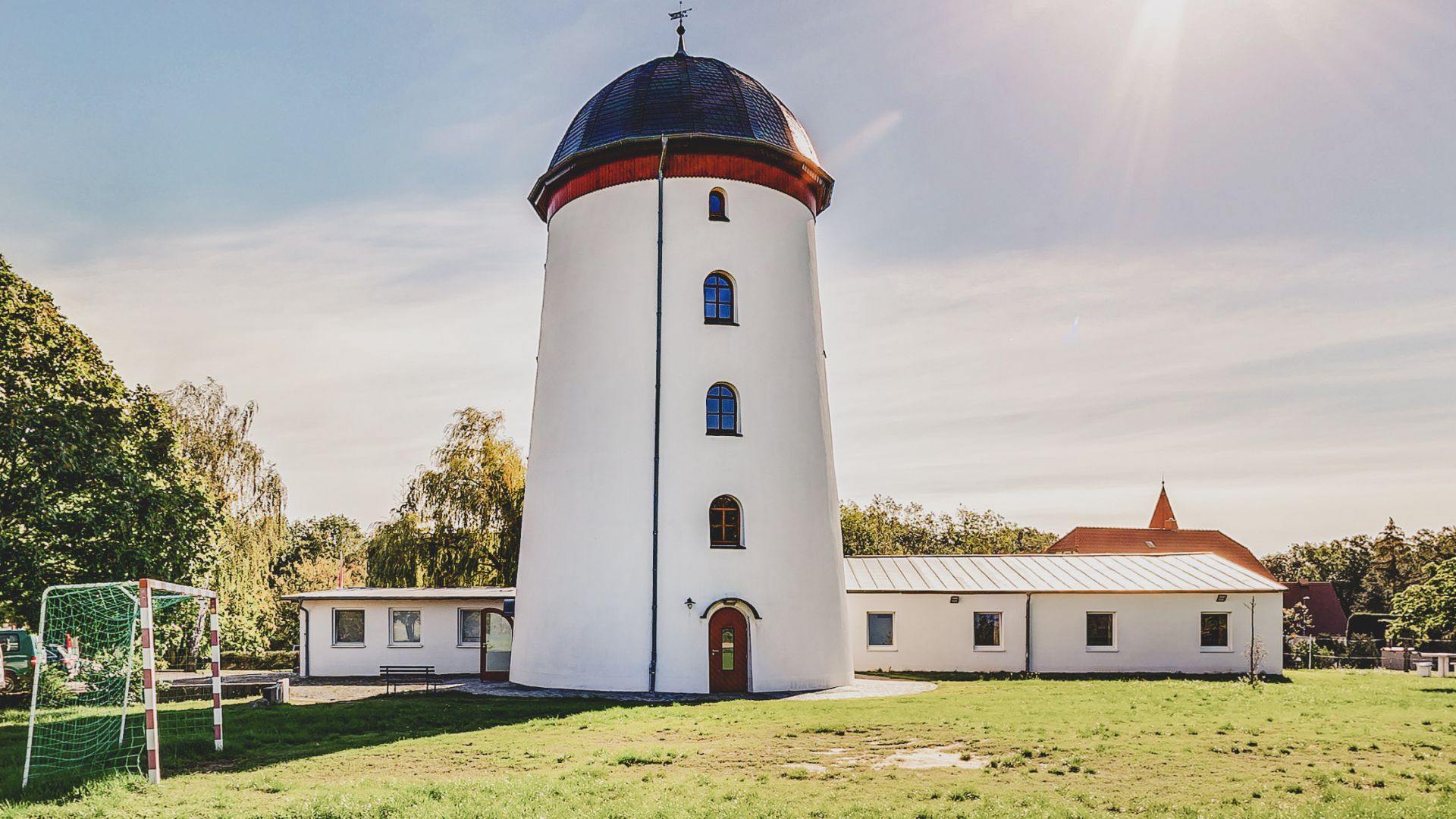Ein weißer Turm mit einem blauen Kuppeldach steht zentral in einem begrünten Bereich. Links im Vordergrund befindet sich ein Fußballtor, dahinter ein flaches Gebäude mit mehreren Fenstern. Im Hintergrund ist eine Kirche mit einem spitzen roten Dach erkennbar. Die Szene ist bei strahlendem Sonnenschein aufgenommen.