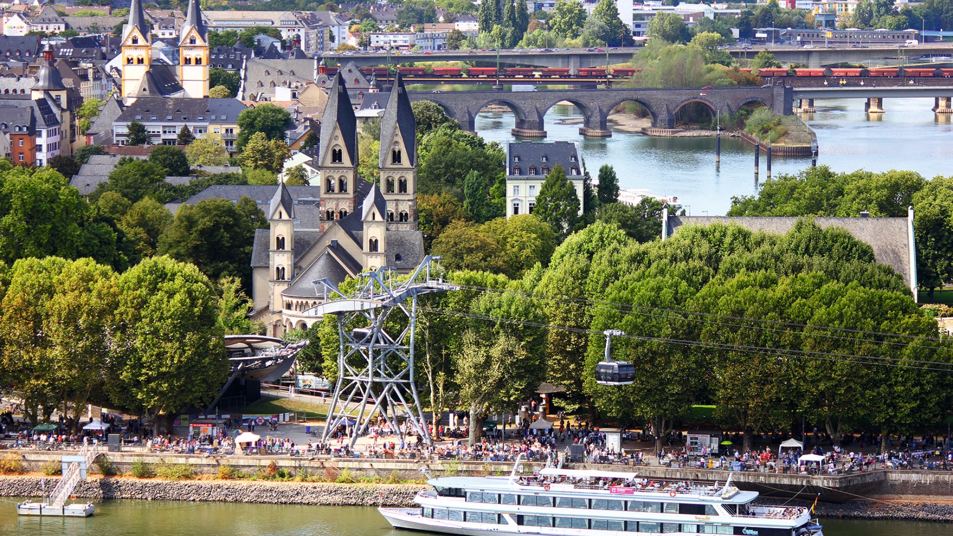 Ein großes Boot fährt auf dem Fluss in Koblenz, während eine Seilbahn über das Wasser fährt. Viele Menschen stehen am Flussufer mit Bäumen. Im Hintergrund befinden sich Kirchen, Häuser, Brücke und weitere Grünflächen.