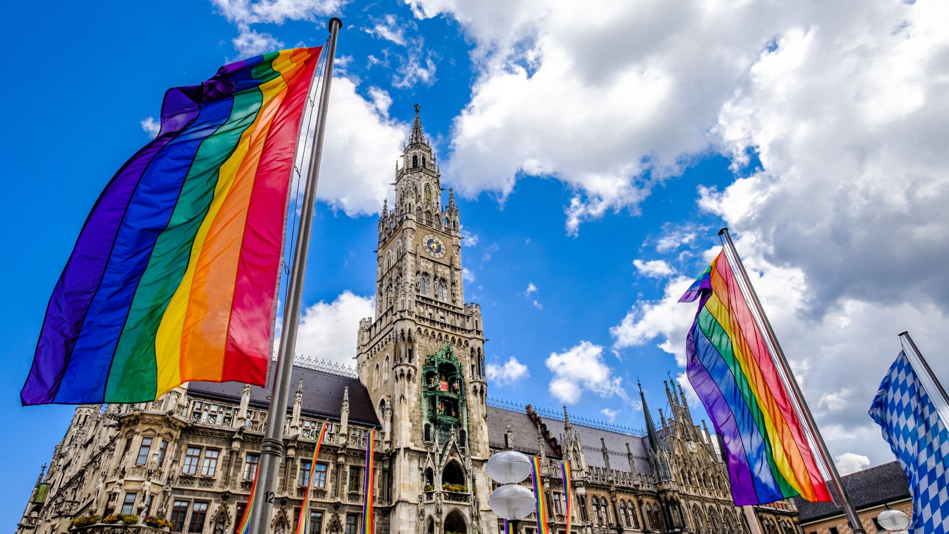 New City Hall in Munich with several rainbow flags in the foreground. The building has a high tower with a clock and is surrounded by blue and white flags.