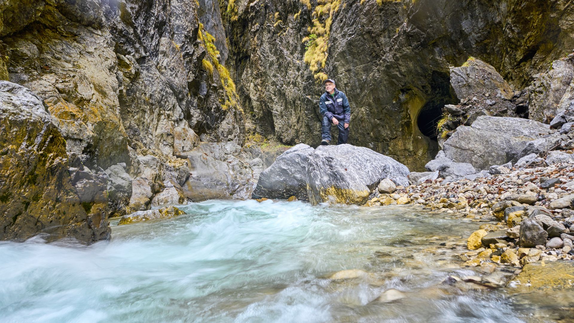 In Garmisch-Partenkirchen steht ein Klammverwalter in der Partnachklamm auf einen großen Felsen. Er ist umgeben von steilen, felsigen Klippen und einem fließenden Bach mit klarem, blauem Wasser.