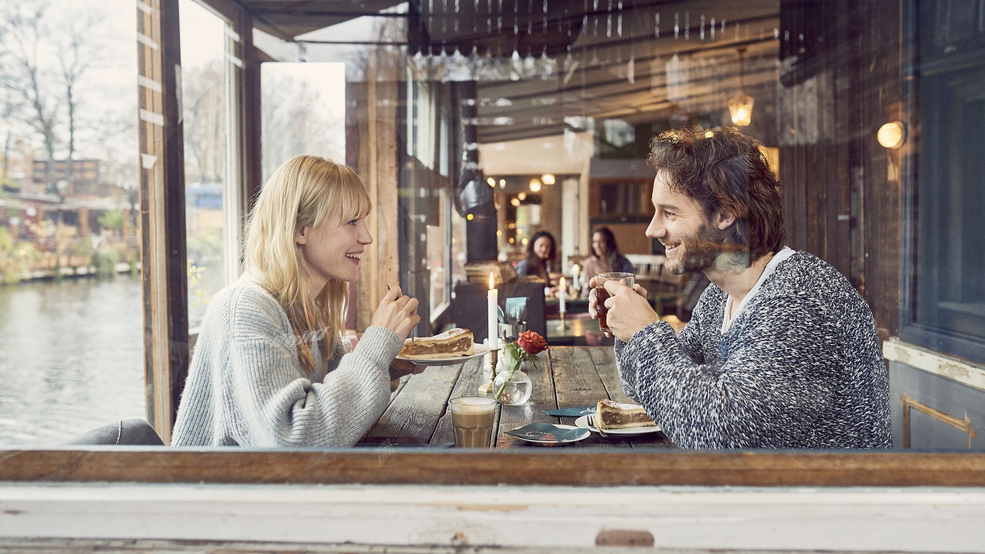 2 Gäste sitzen an einem Holztisch im Restaurant Freischwimmer an der Spree in Kreuzberg. Sie genießen Kaffee und Kuchen und unterhalten sich miteinander.