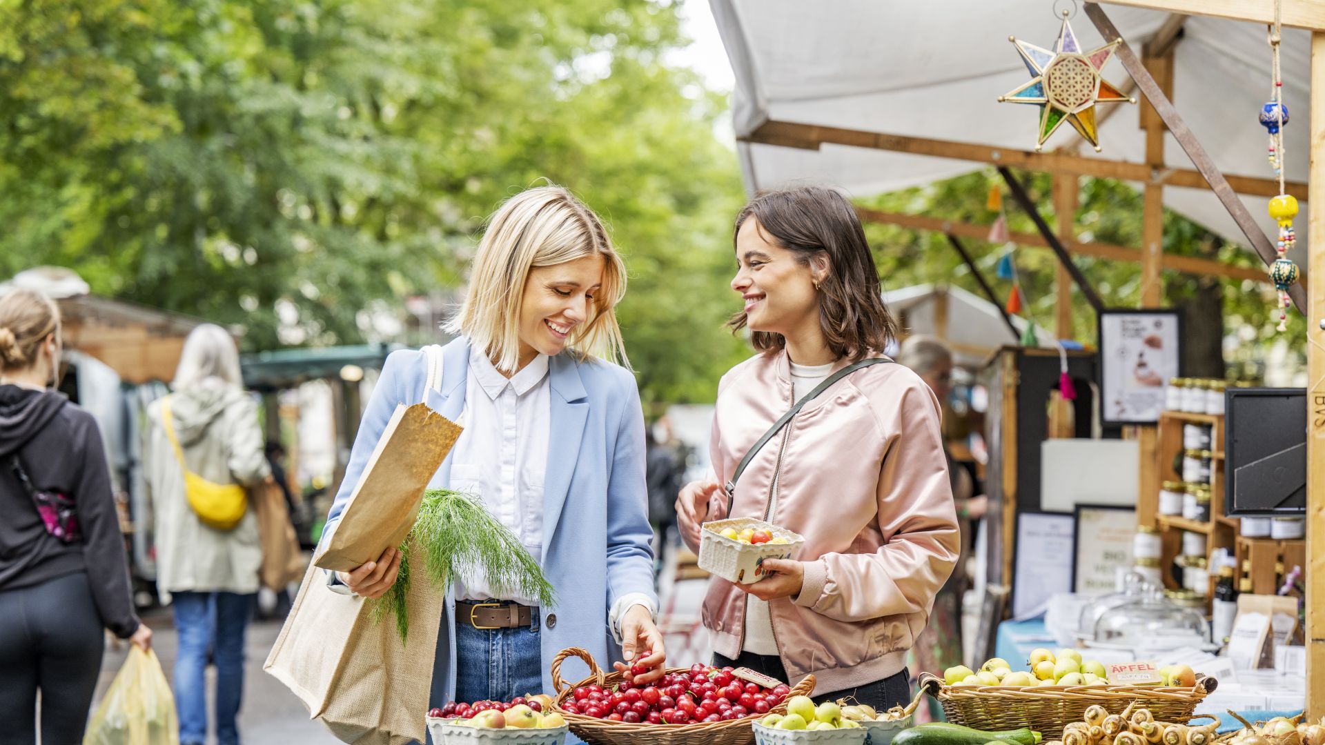 Zwei Personen stehen an einem Marktstand am Kollwitzplatz in Berlin. Sie sehen sich frische Produkte an, darunter einen Korb mit Kirschen, um den herum weiteres Obst und Gemüse liegt. Eine Person hat eine Tasche mit Grünzeug um den Arm, während die andere ein Körbchen mit Obst in der Hand hält. Im Hintergrund sind Marktstände und andere Besucher unter den Bäumen zu sehen.