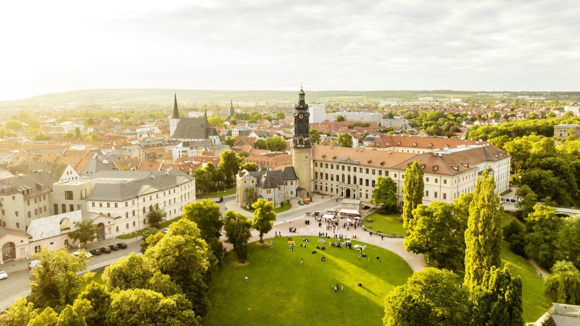 Die Luftaufnahme von Weimar in Thüringen zeigt ein historisches Gebäude mit einem hohen Uhrenturm in der Mitte. Rot gedeckte Gebäude und Kirchen mit spitzen Türmen sind über das Gebiet verteilt. Im Vordergrund ist ein großer grüner Park mit Bäumen und auf dem Rasen versammelten Menschen zu sehen. Der Himmel ist teilweise bewölkt.