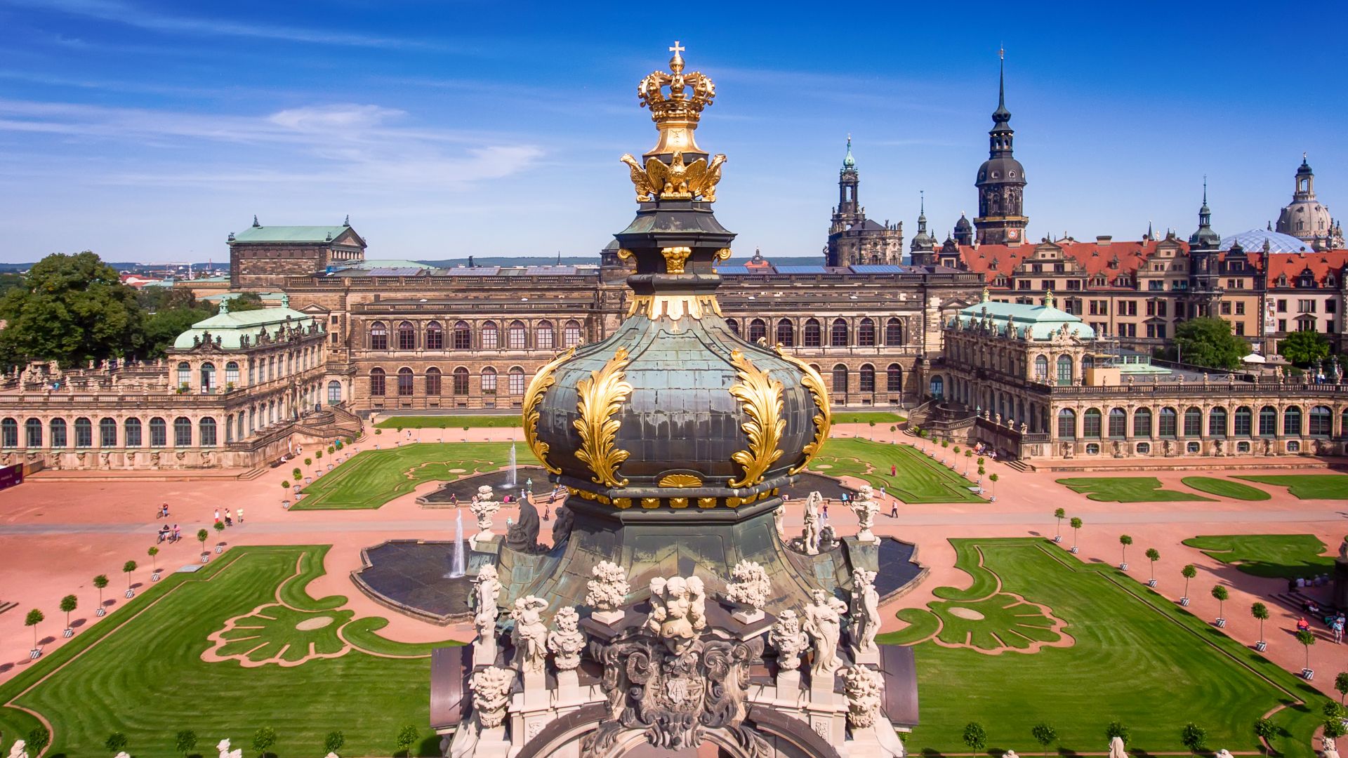 Eine große, verzierte Kuppel der Dresdner Zwinger mit goldenen Details steht im Vordergrund. Im Hintergrund ist ein langes, rechteckiges Gebäude mit gepflegten Rasenflächen.