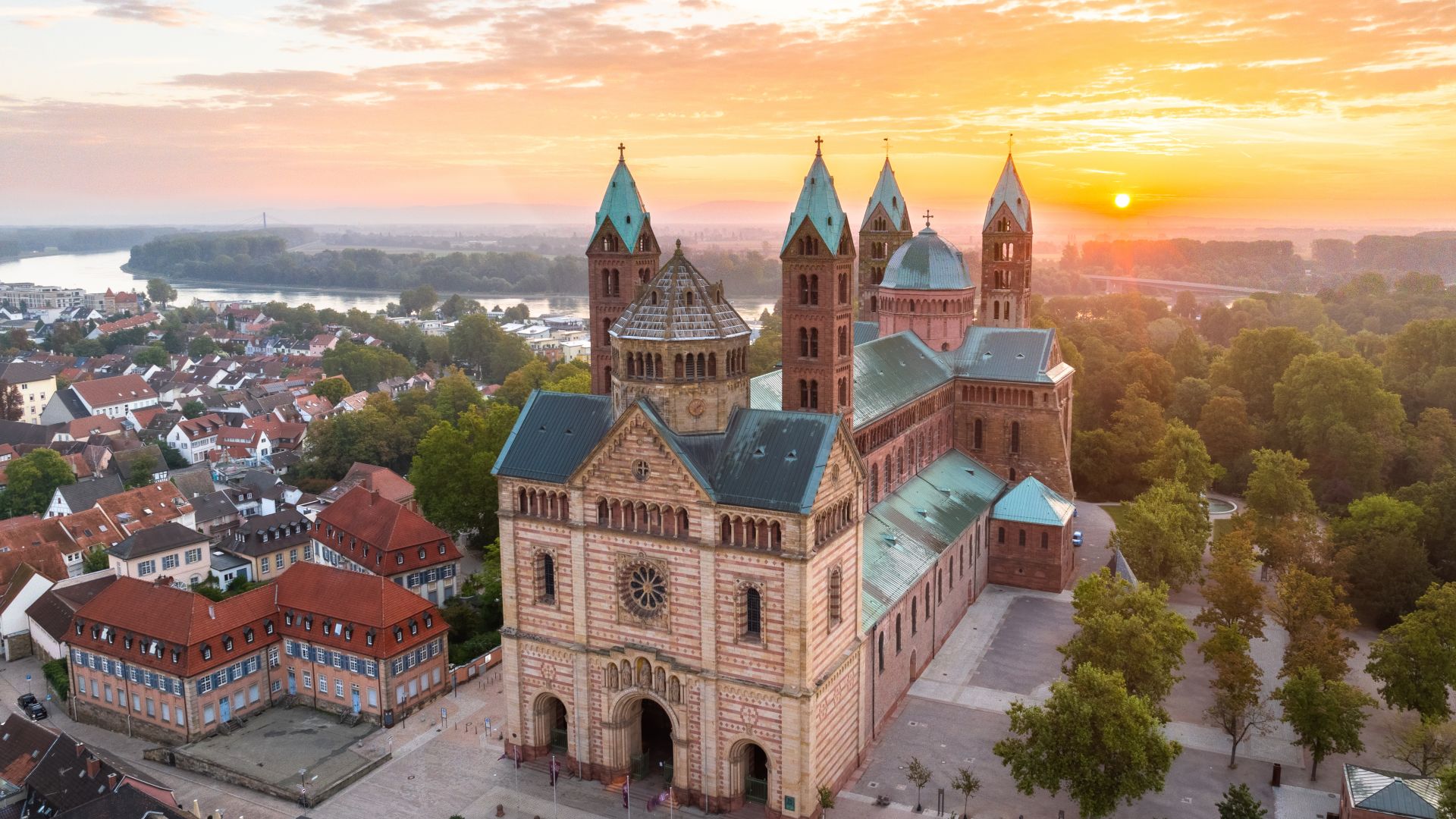 Luftaufnahme des Speyerer Doms in Speyer bei Sonnenaufgang. Der Dom mit grünen Dächern und mehreren Türme, steht prominent im Vordergrund. Kleine Häuser mit roten Dächern sind um den Dom herum gruppiert.